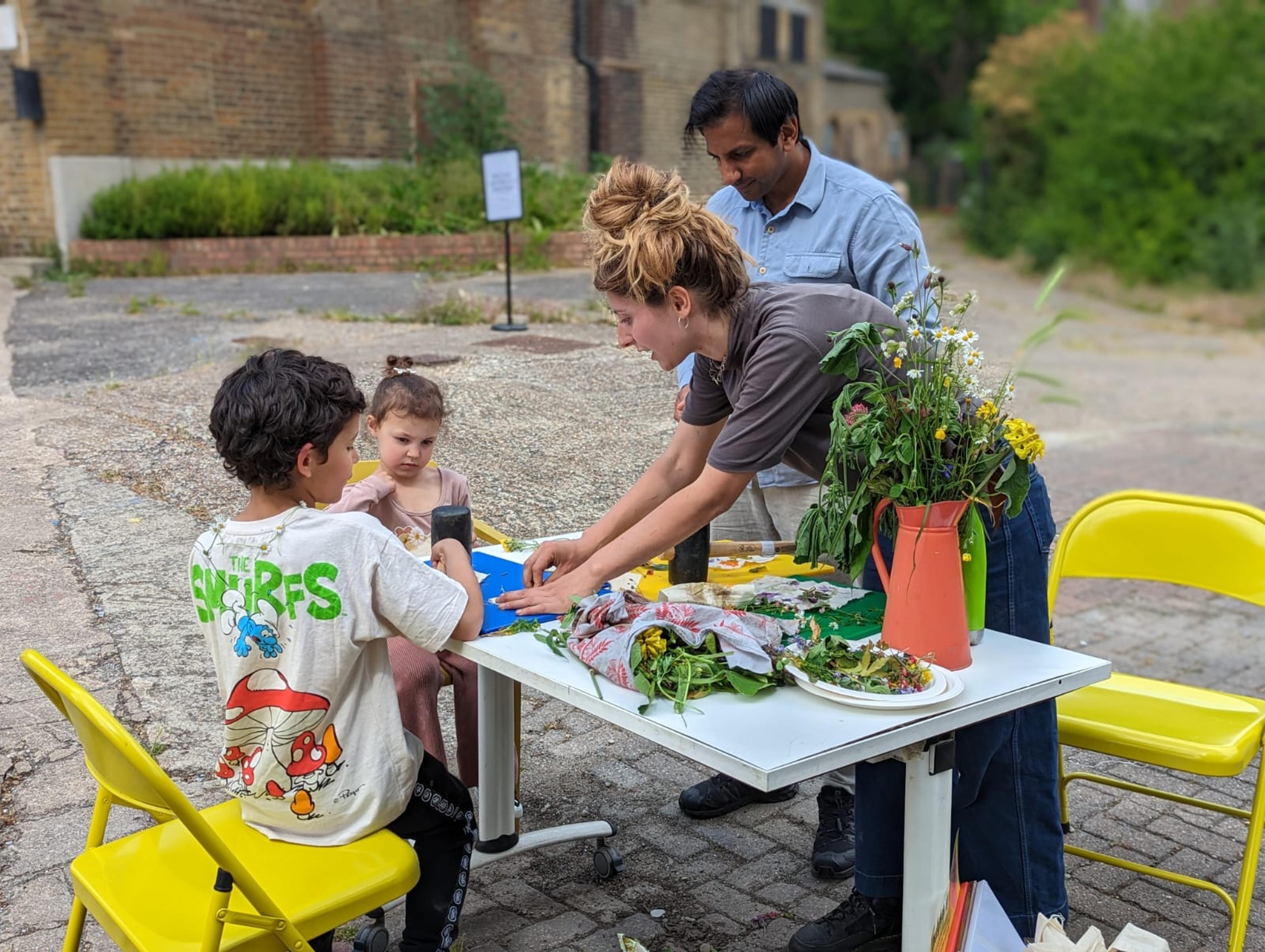 Two adults and two children outside, sitting around a table creating illustrations with flowers. There is a vase of flowers on the table.