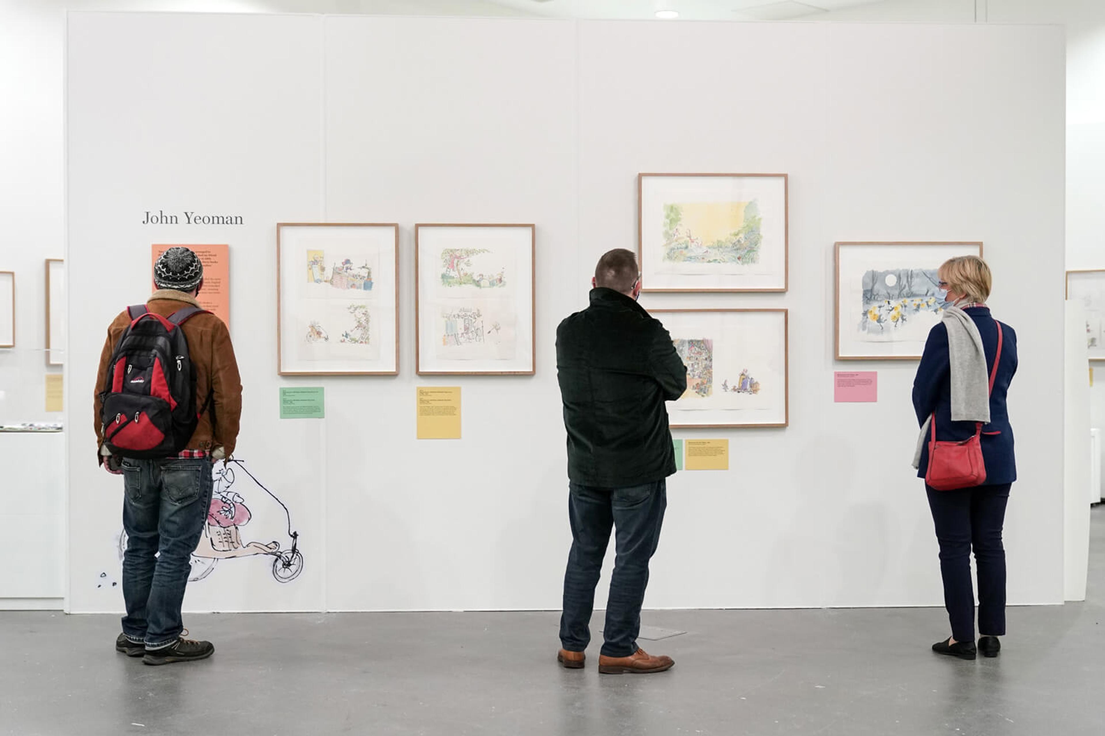 Three people in an exhibition space with framed illustrations on the wall.