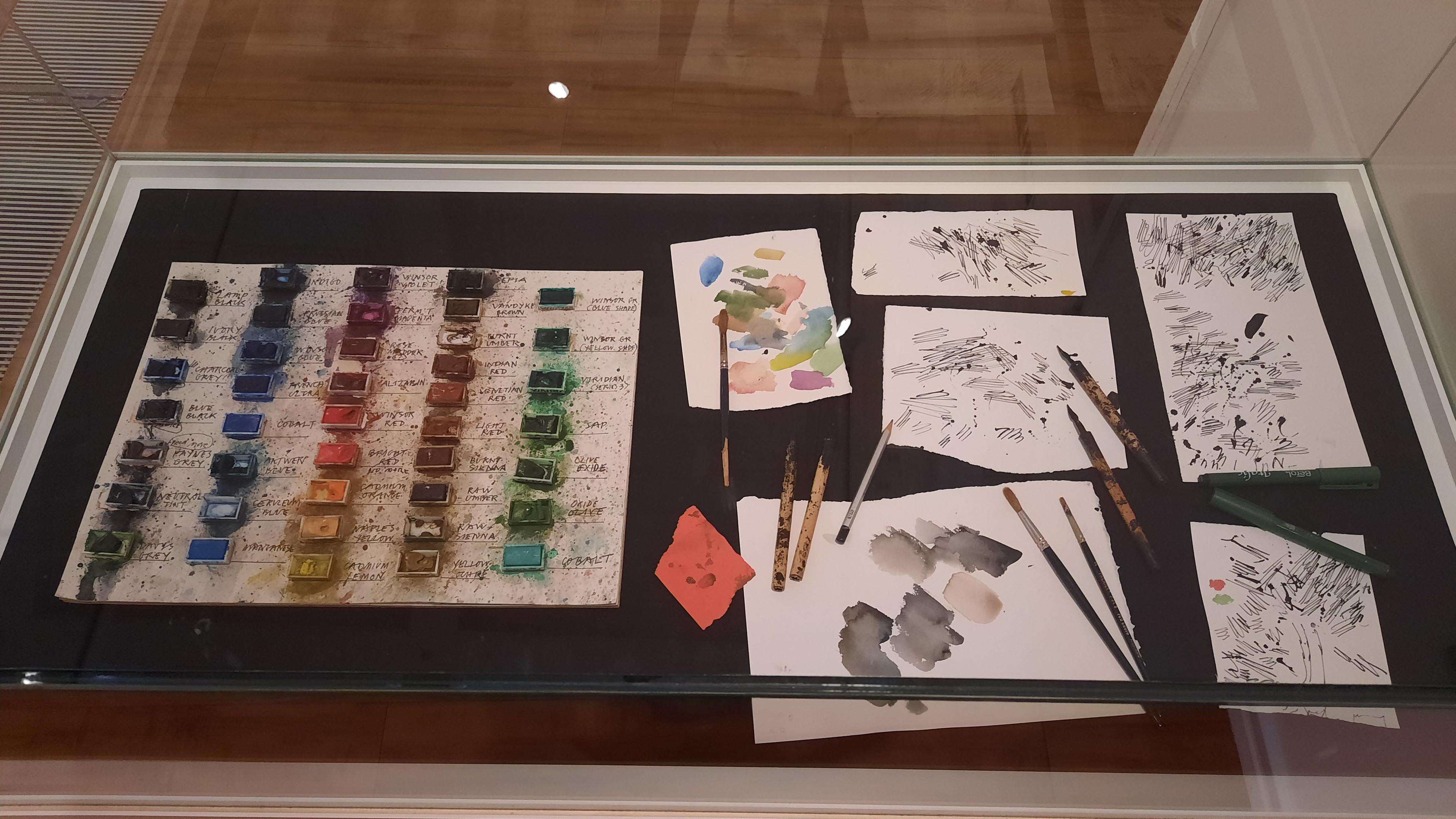 Paints, brushes and pieces of paper with test marks on them in a glass cabinet.