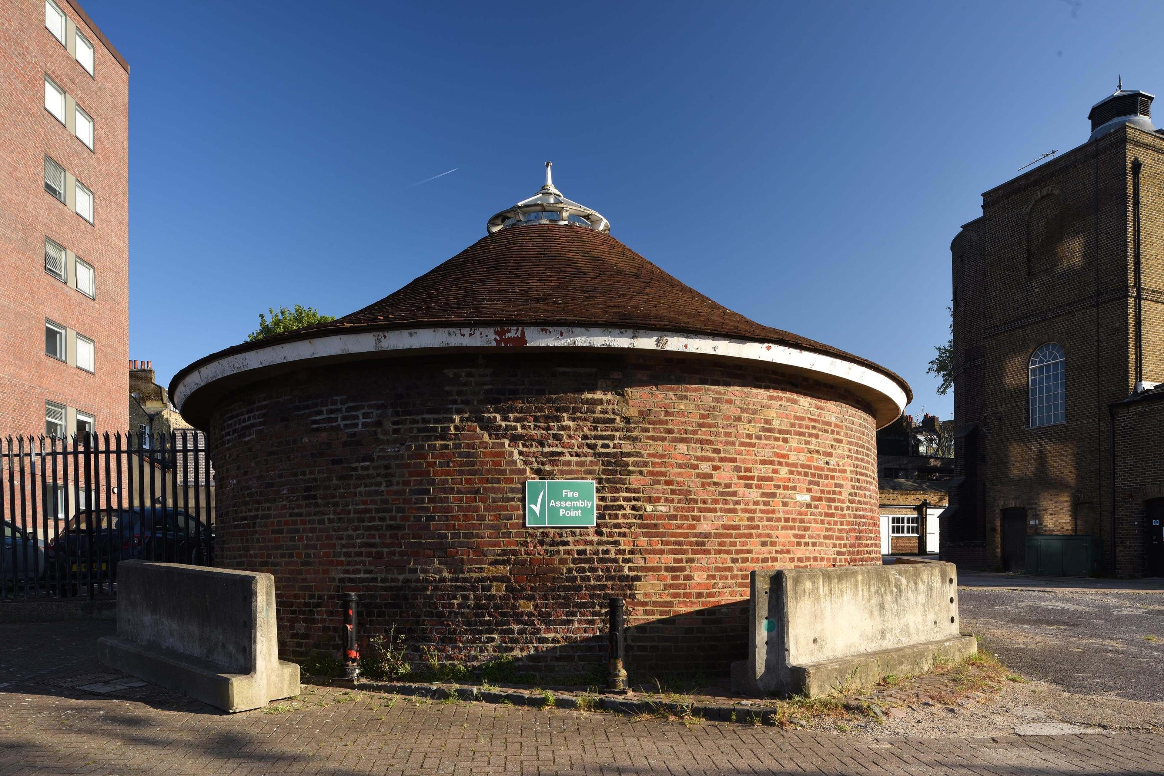 Photograph of a round brick building with a block of flats on the left and a large brick building on the right, with a blue sky in the background.