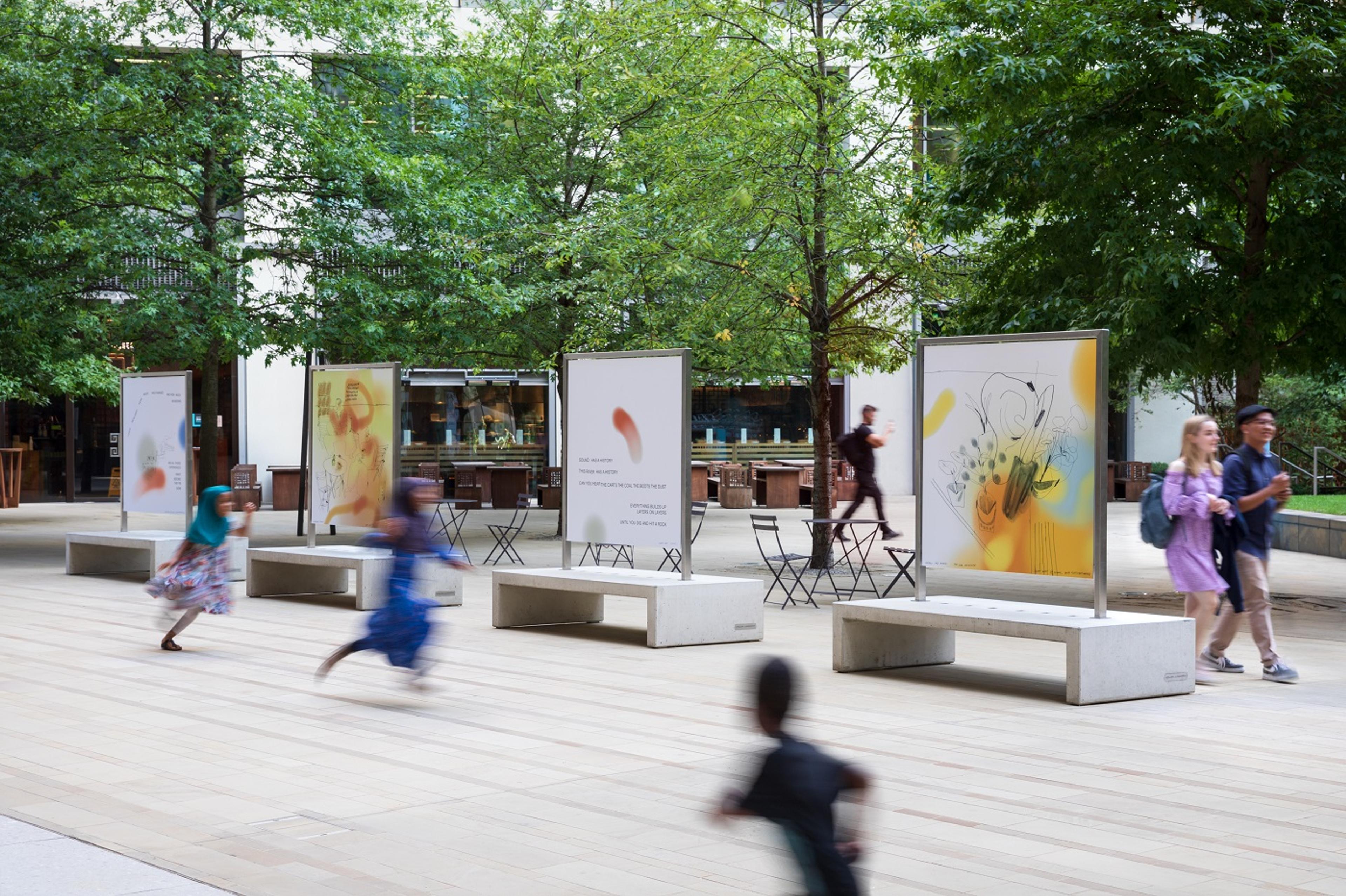 Freestanding large scale prints installed outdoors with trees in the background and children running in the foreground