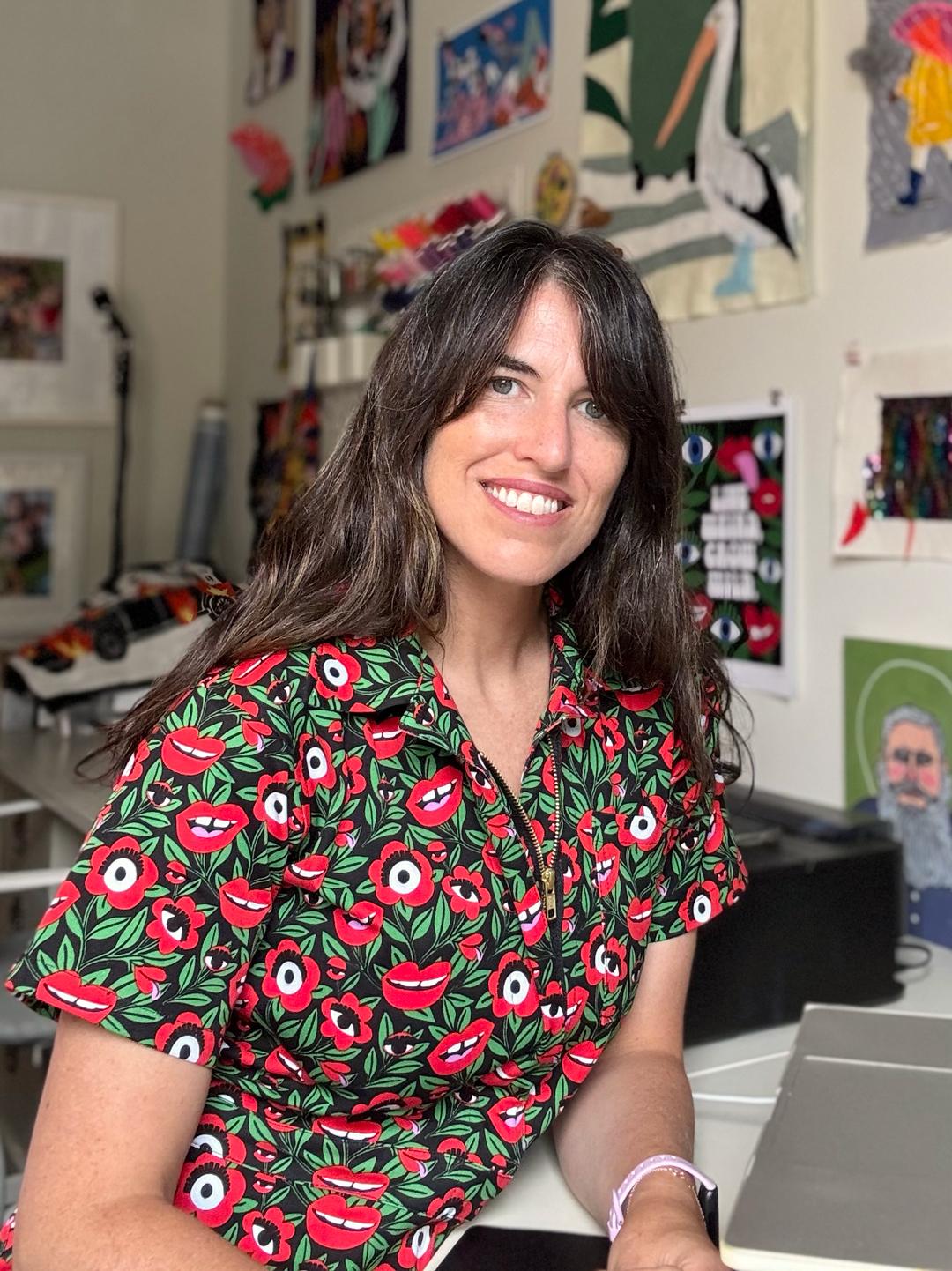 A woman with brown hair smiles, she wears a patterned top and is in a studio with images on the wall. 