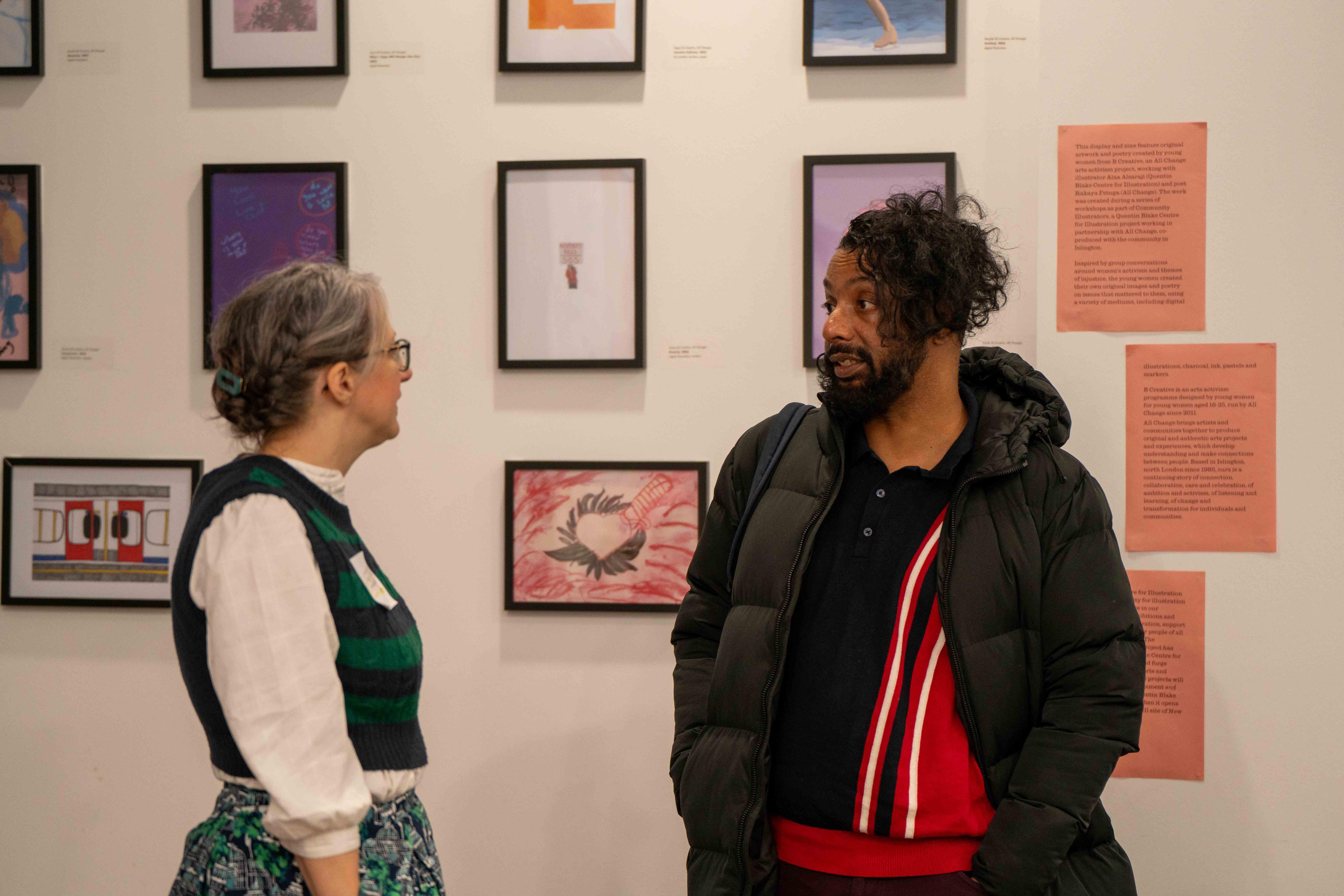 Two people stand and chat, framed illustrations are on the wall in the background.