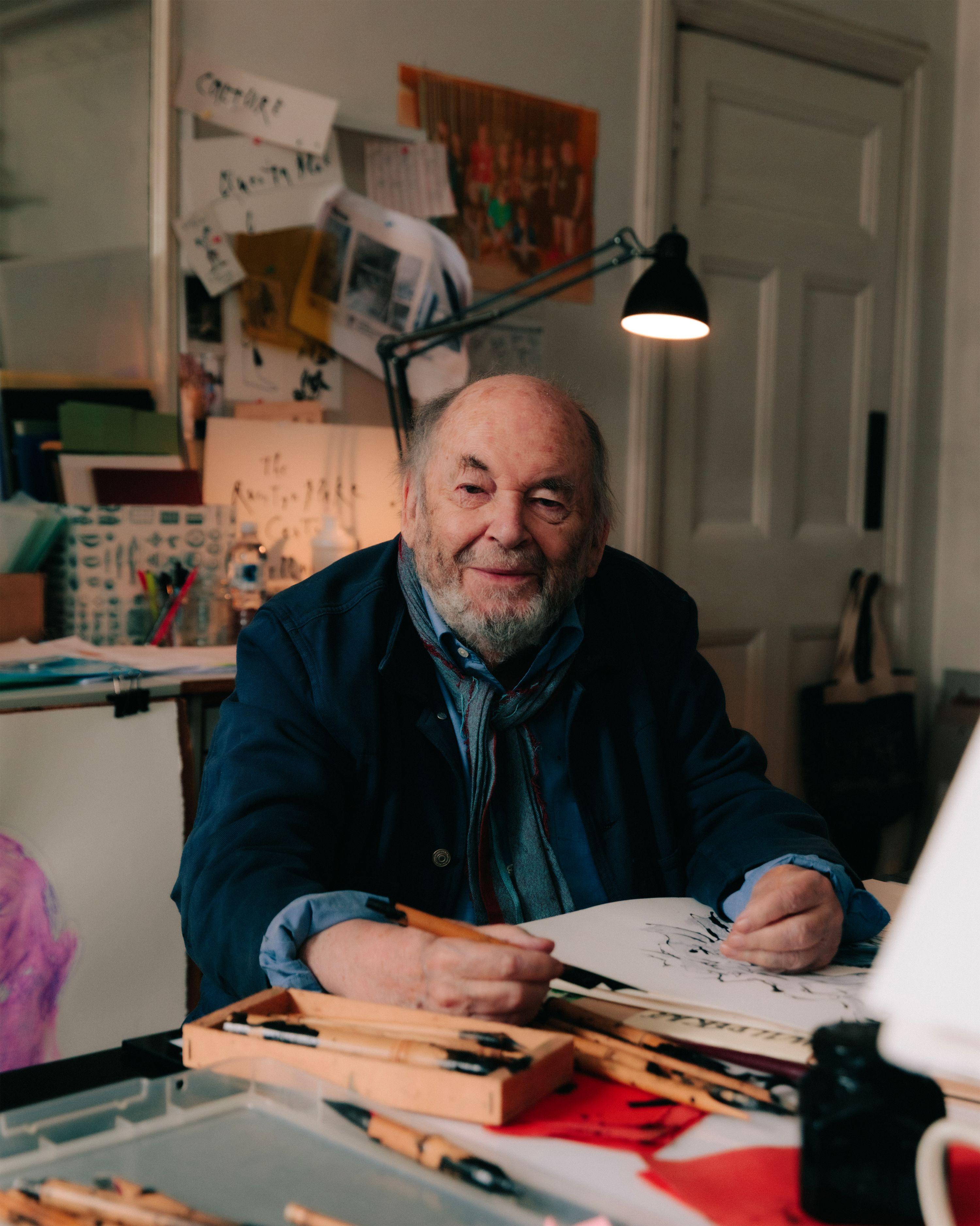 A friendly bearded man man smiling while creating a pen and ink drawing in a studio filled with artwork and photographs.