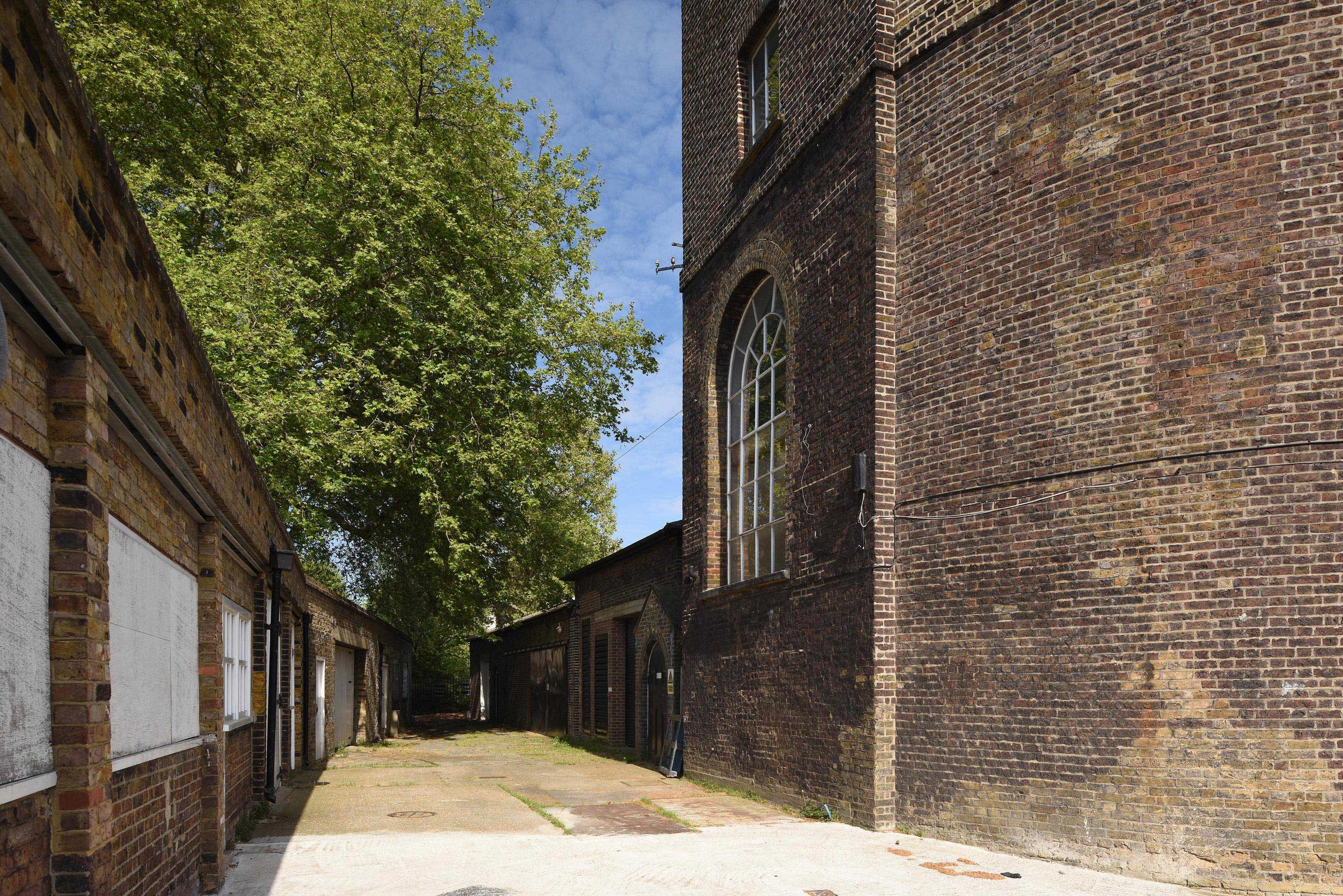 Photograph of a cobbled path with a tree at one end and two brick buildings either side. The sky is blue.