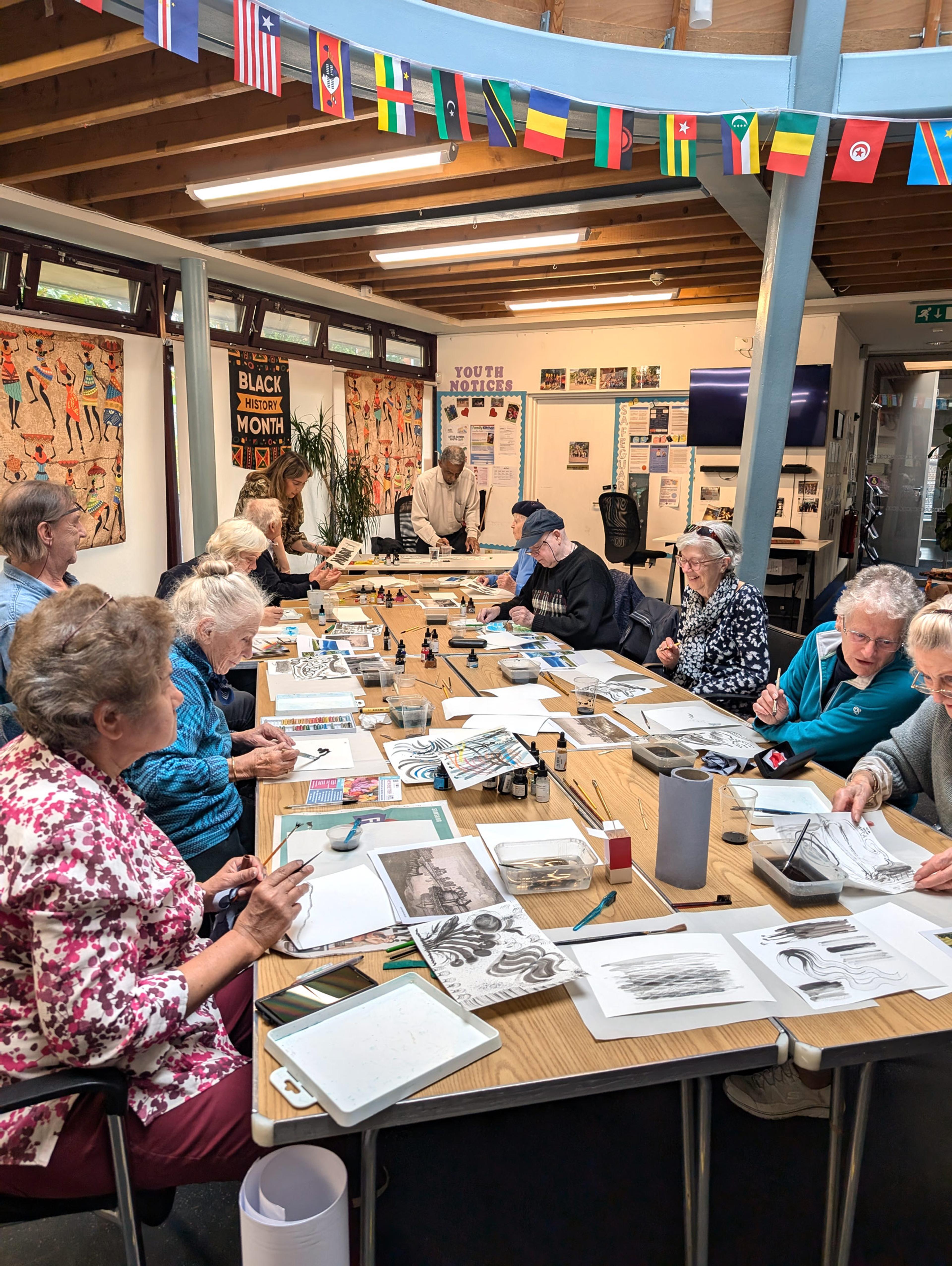 A group of older adults gathered around a large table. They are painting with diluted ink on sheets of paper. 