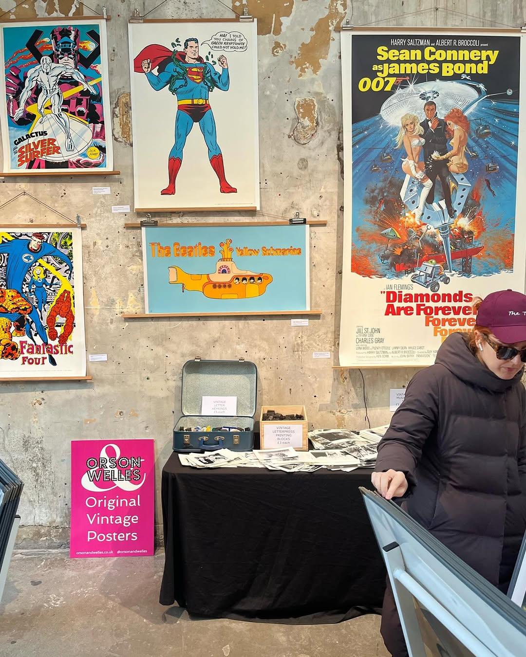 Vintage poster display on a textured wall, featuring Superman, James Bond, The Beatles, and The Fantastic Four. A person browses at a stand.