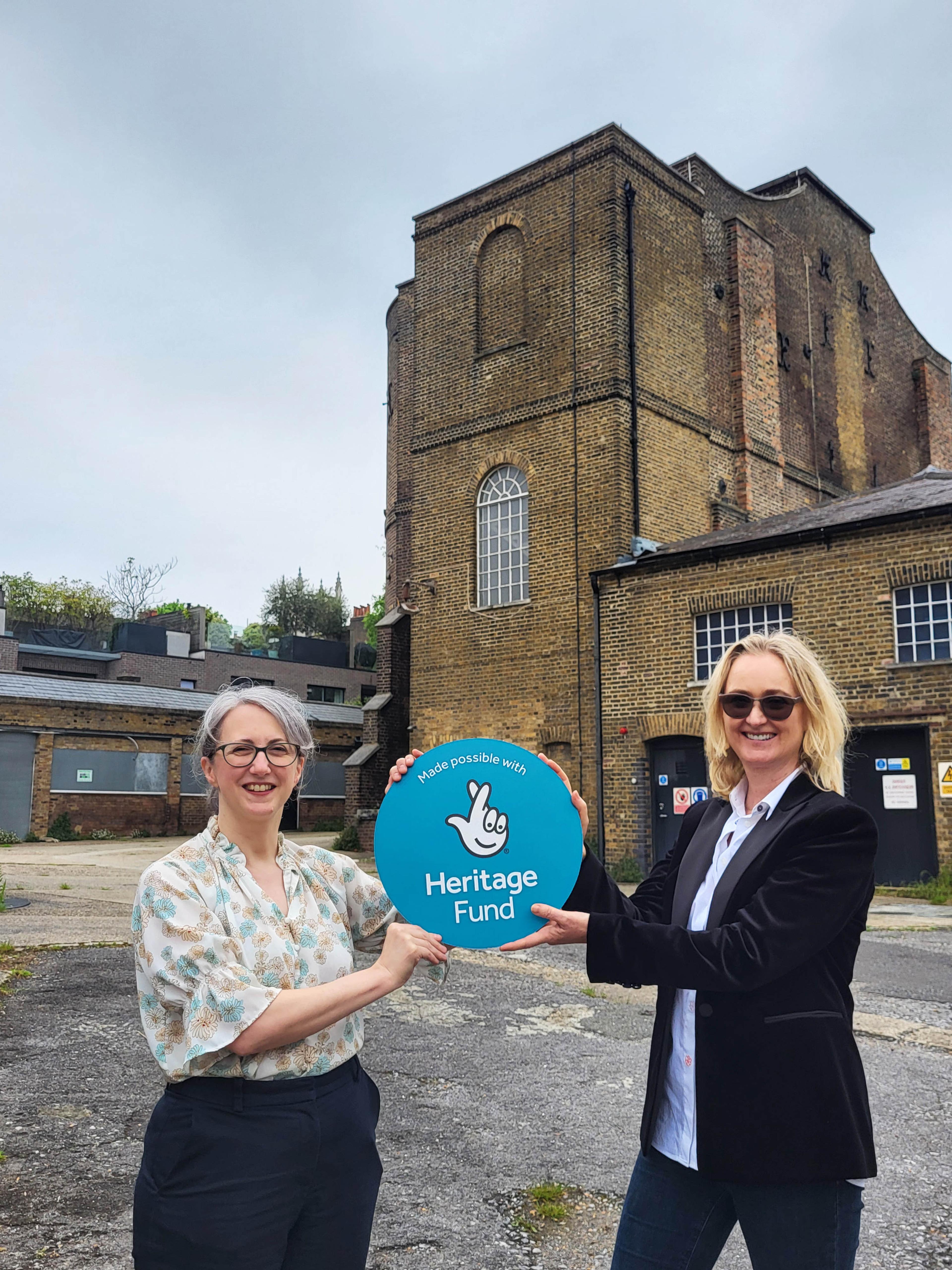 Photograph of two people standing and holding a sign between them with The National Lottery Heritage Fund logo on a teal background, behind them is a brick building.