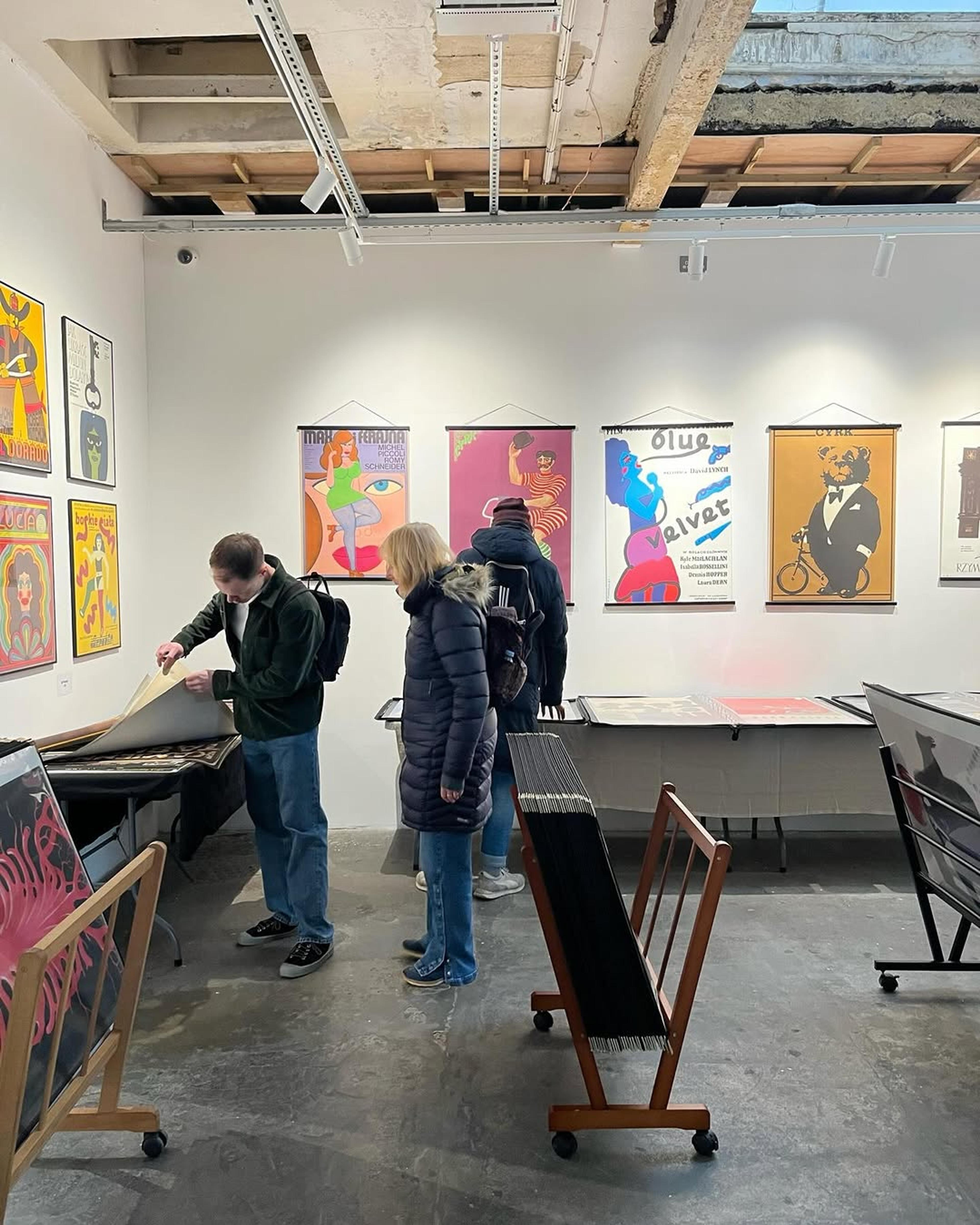 Three people in a gallery examine vintage posters on display. Vibrant retro artwork hangs on white walls