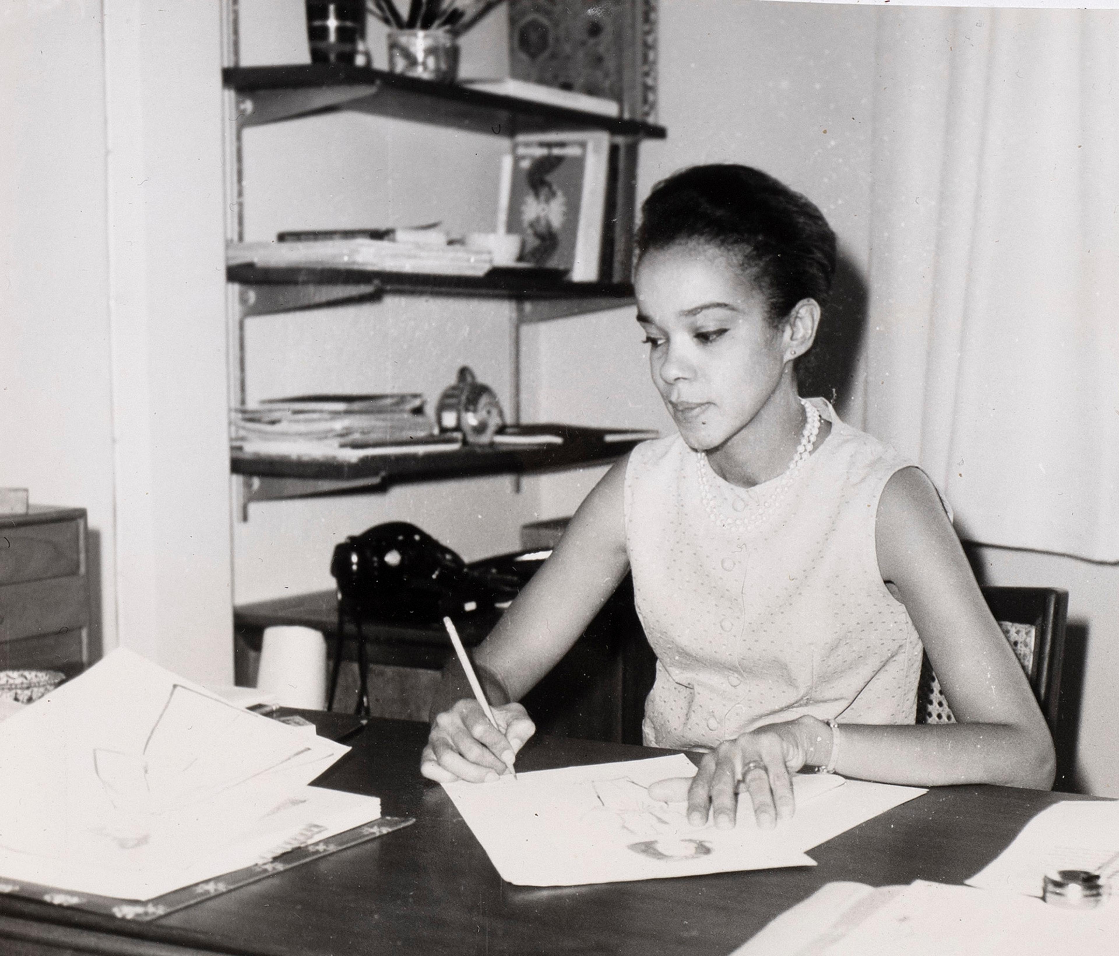 Black and white photograph of a young Black woman with her hair up, drawing at a desk