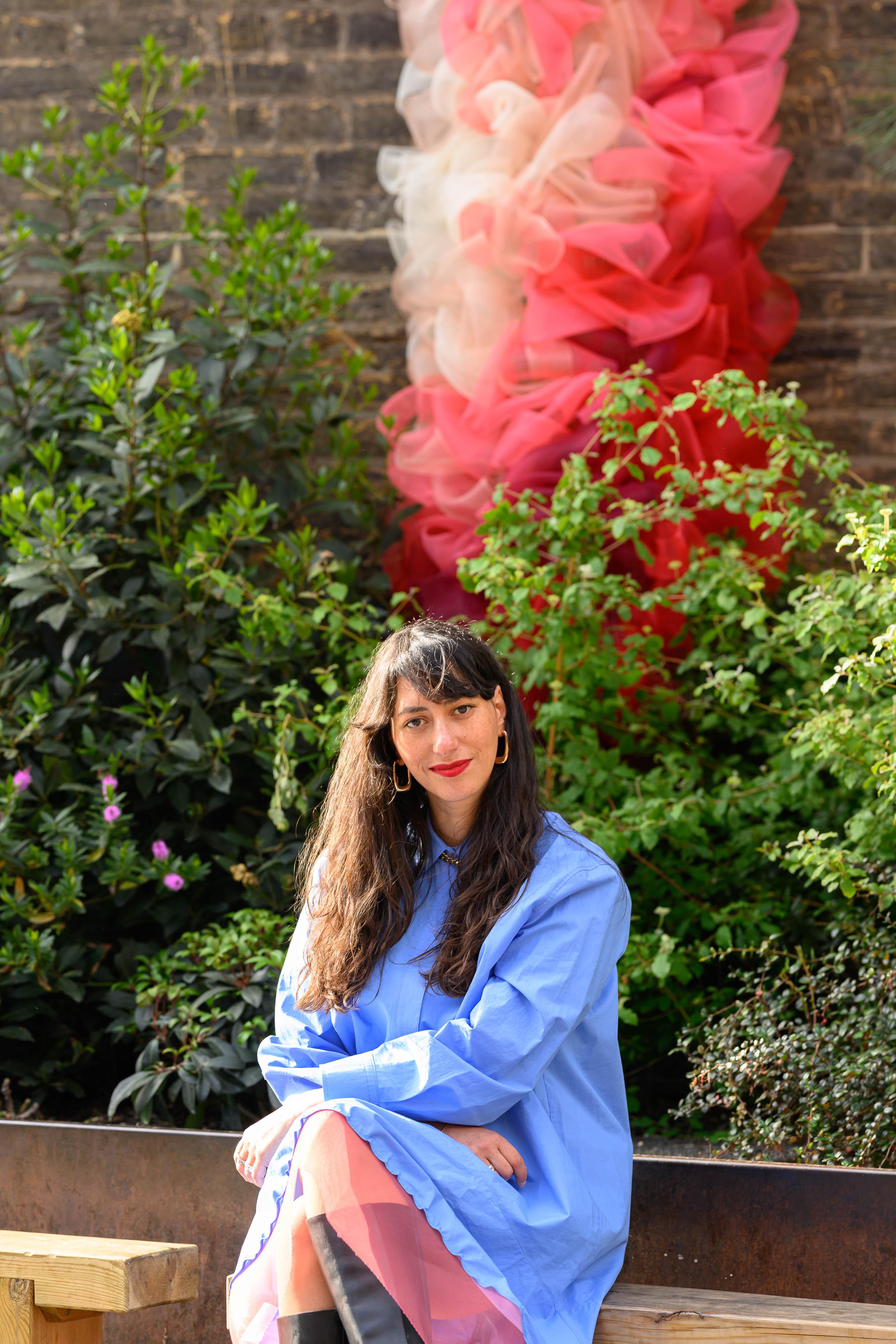 An artist in a blue shirt sits looking out to camera, with a public art sculpture behind her.