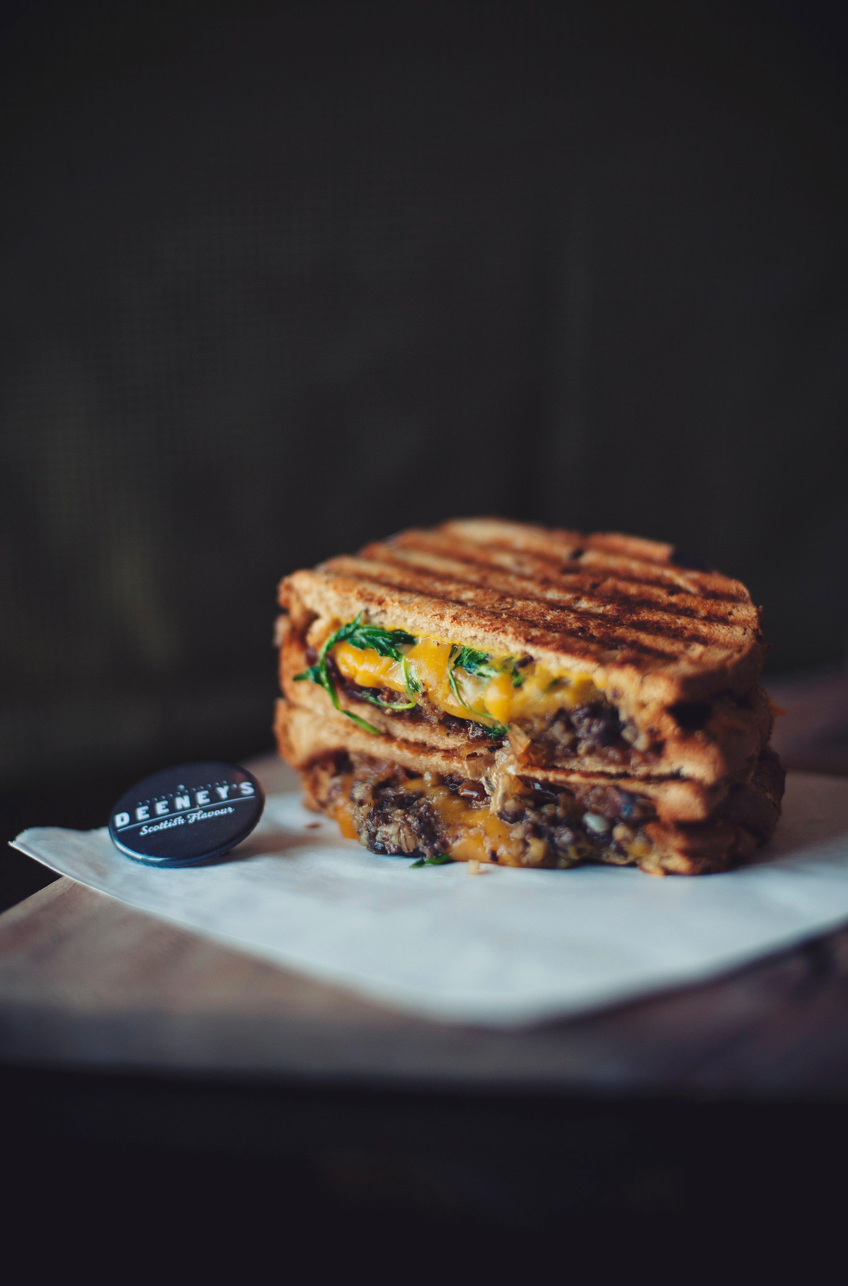 A grilled toastie with melted cheese and haggis oozing out. A pin badge labelled Deeney's is laid beside it. 