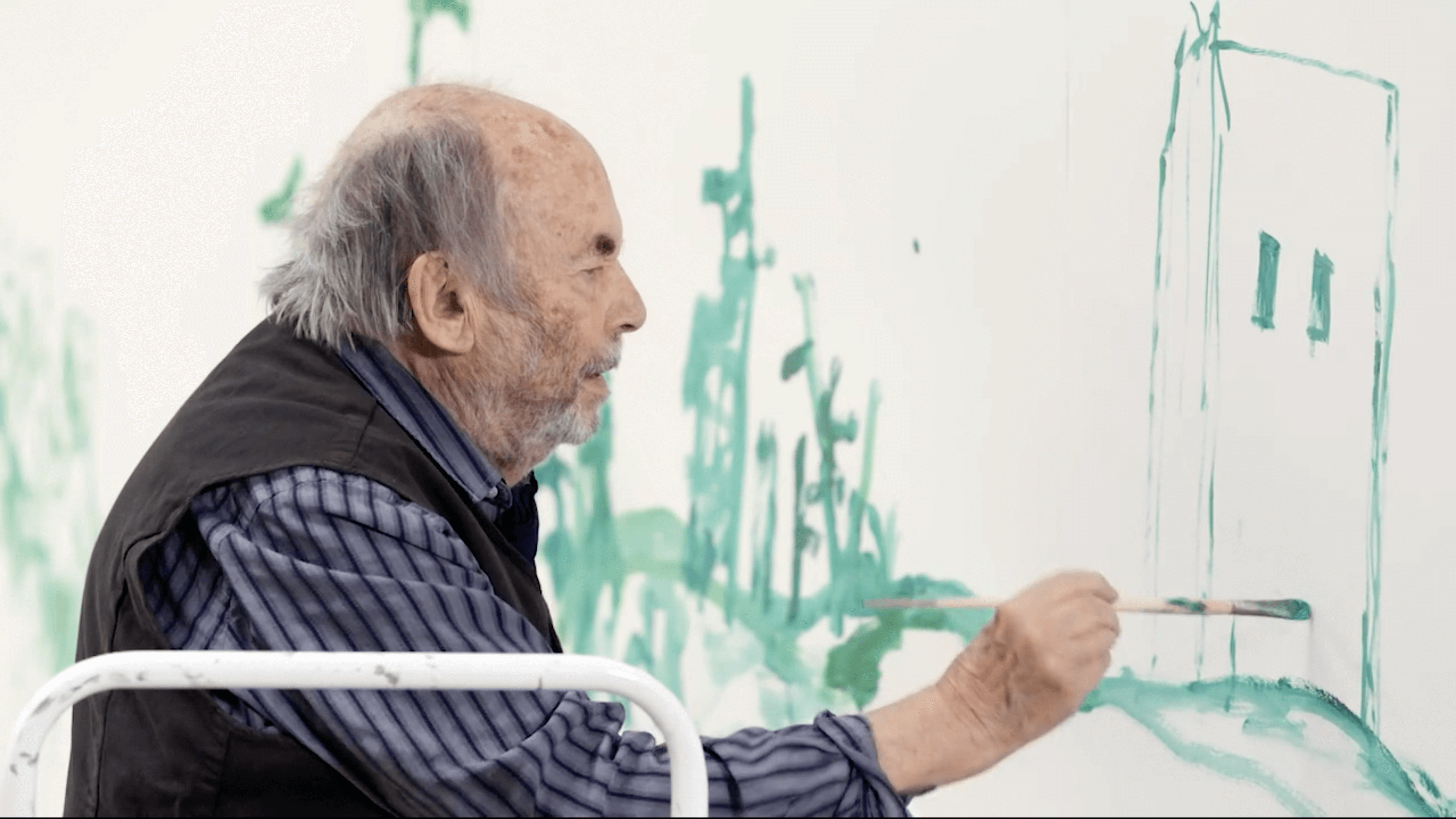 A photograph of Quentin Blake with a paint brush in his hand painting a building in green onto the wall.