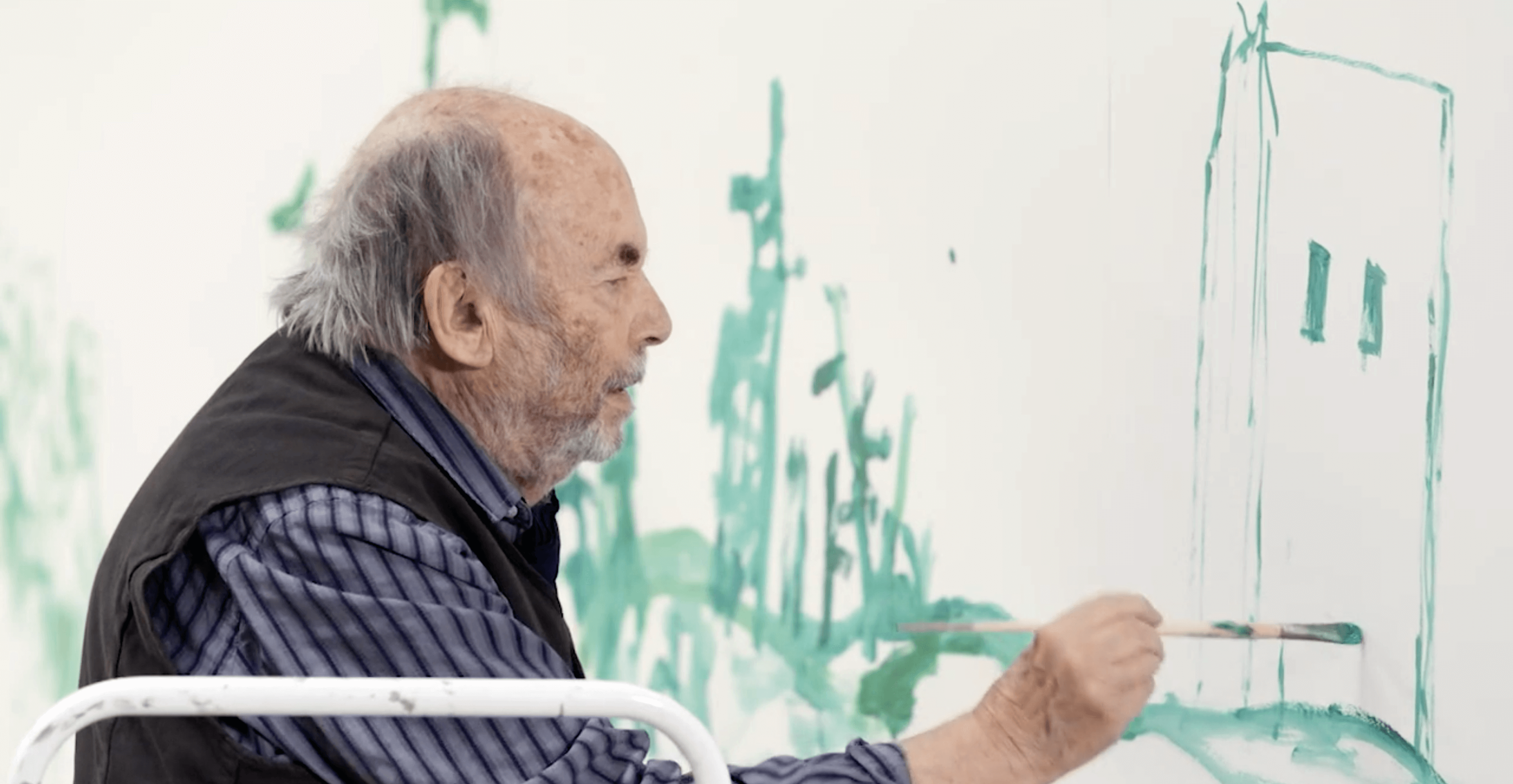 A photograph of Quentin Blake with a paint brush in his hand painting a building in green onto the wall.