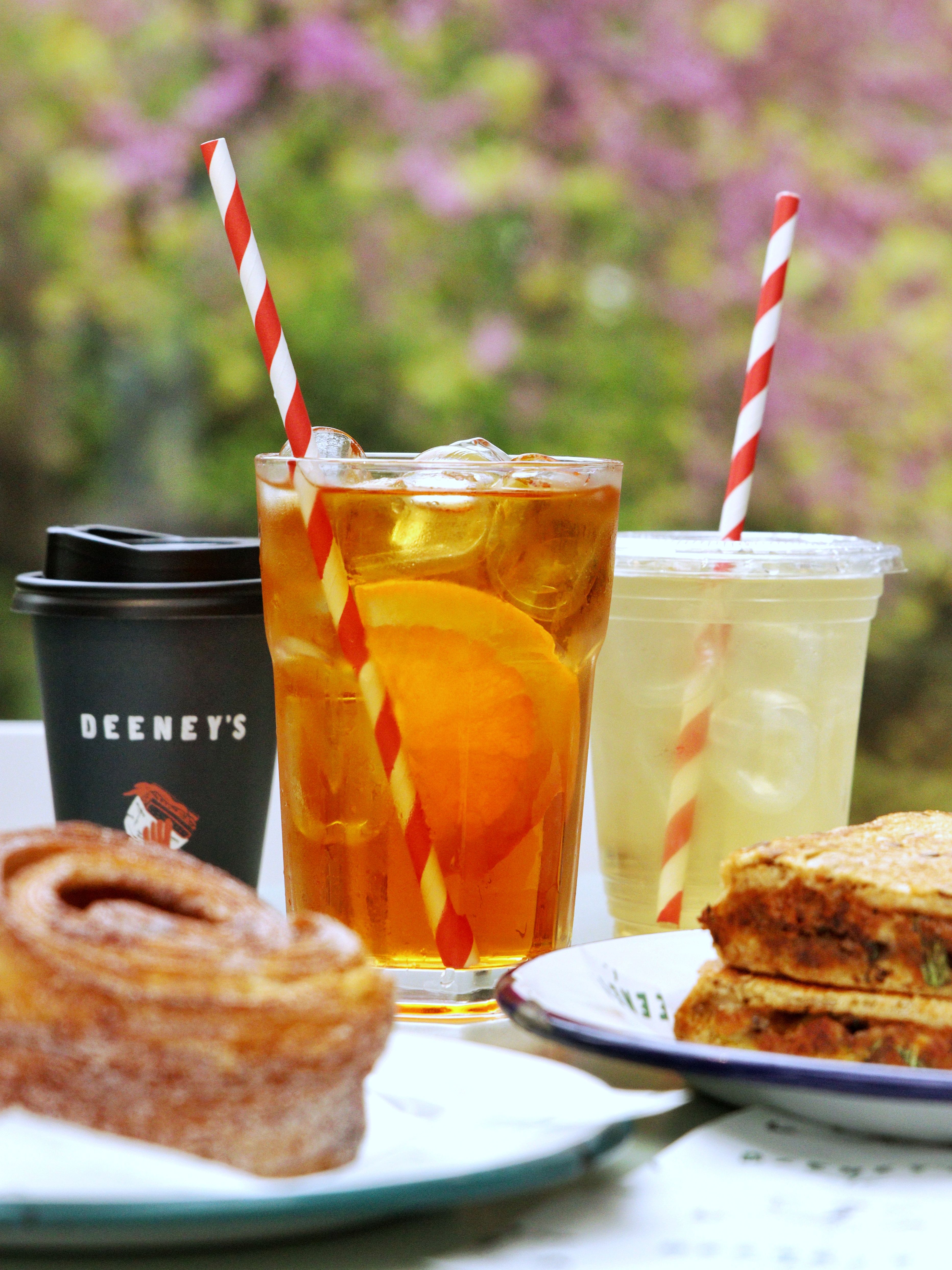 A takeaway coffee cup labelled Deeney's and two iced drinks on a table in the sun. In front, a cinnamon bun and a generously-filled toastie.