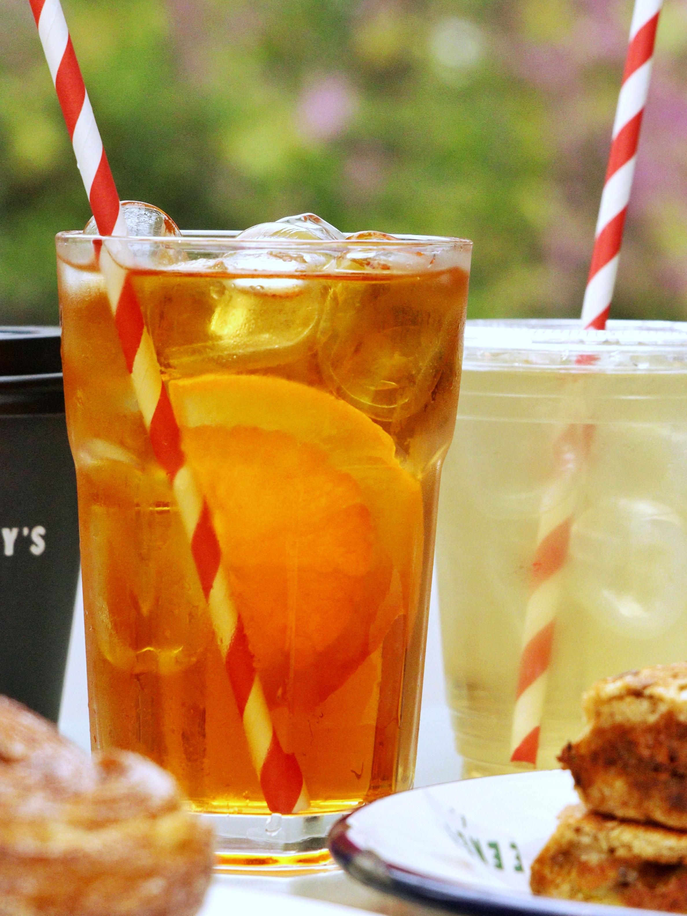 A takeaway coffee cup labelled Deeney's and two iced drinks on a table in the sun. In front, a cinnamon bun and a generously-filled toastie.