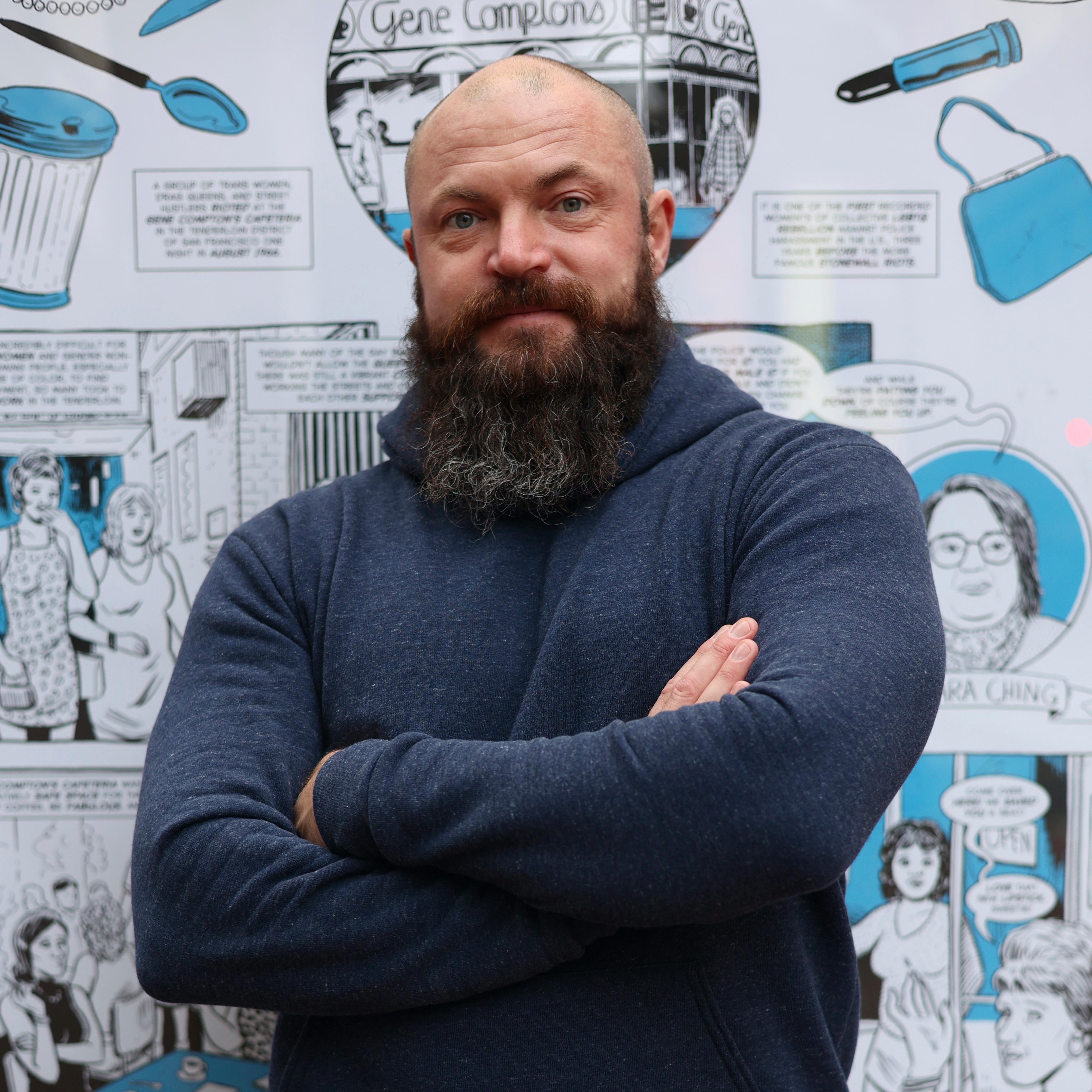 A man stands looking out to camera with his arms folded. Behind him are comic panels.