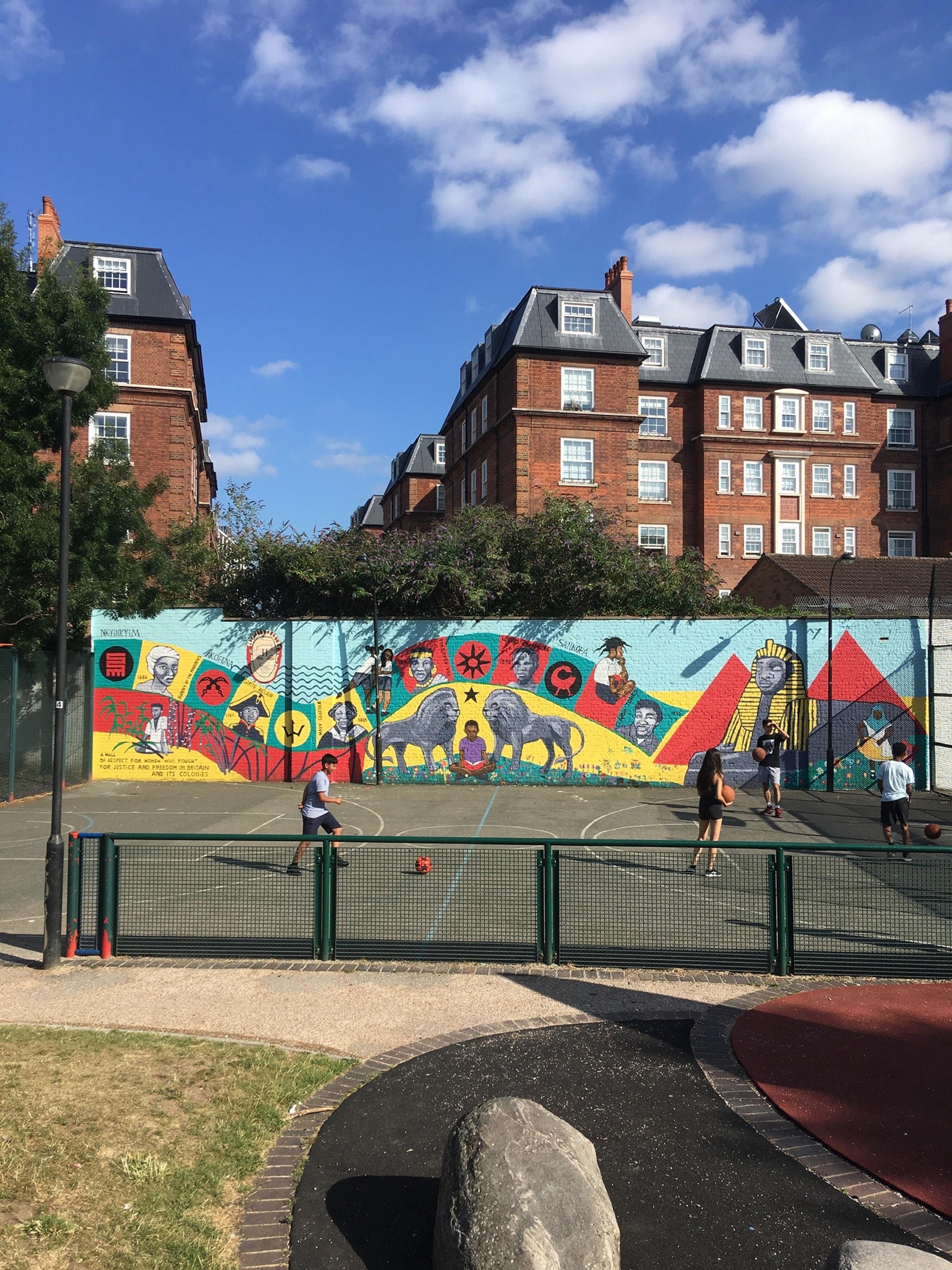 Photograph of a people playing ball games in a park with a mural on the wall in the background. There are tall buildings in the background.