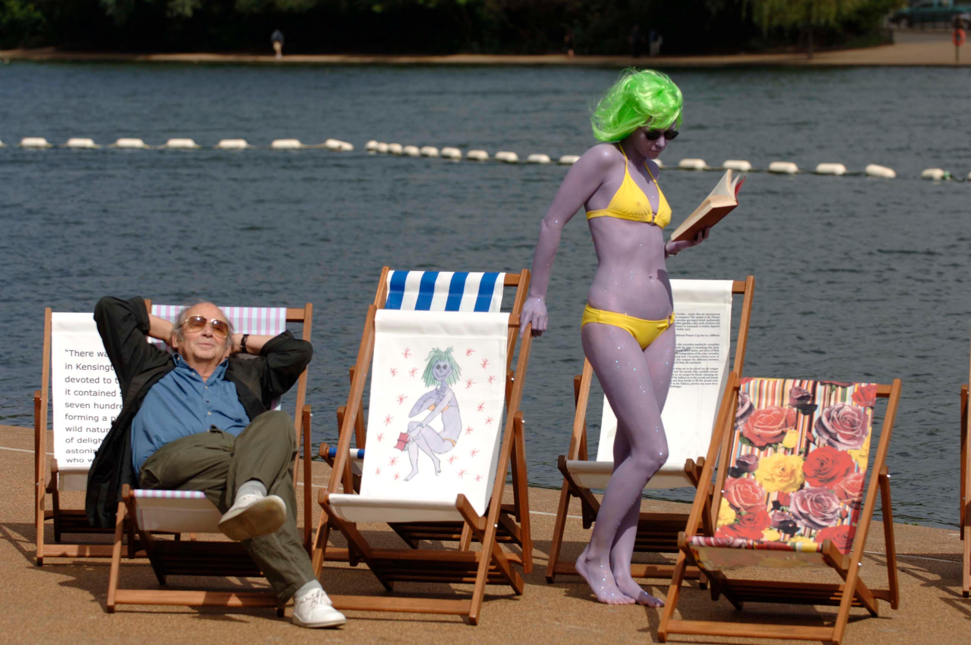A photograph of a man in sunglasses reclining in a deckchair. A woman with green hair, purple skin and a yellow bikini is standing next to him. There is an empty deckchair next to them which has an illustration of the same woman printed on it.