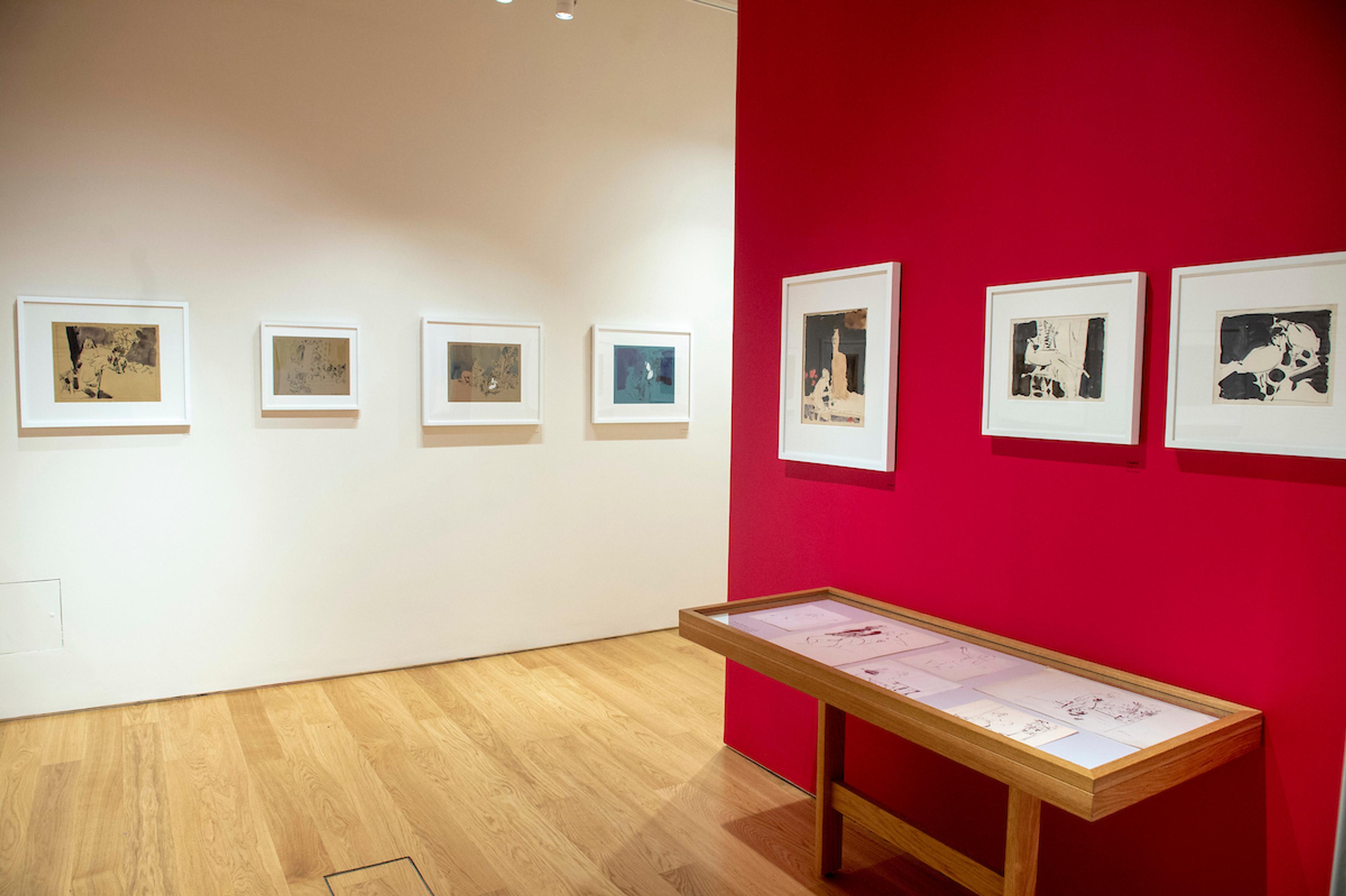 Framed illustrations displayed on gallery walls with a glass display cabinet in an exhibition space.