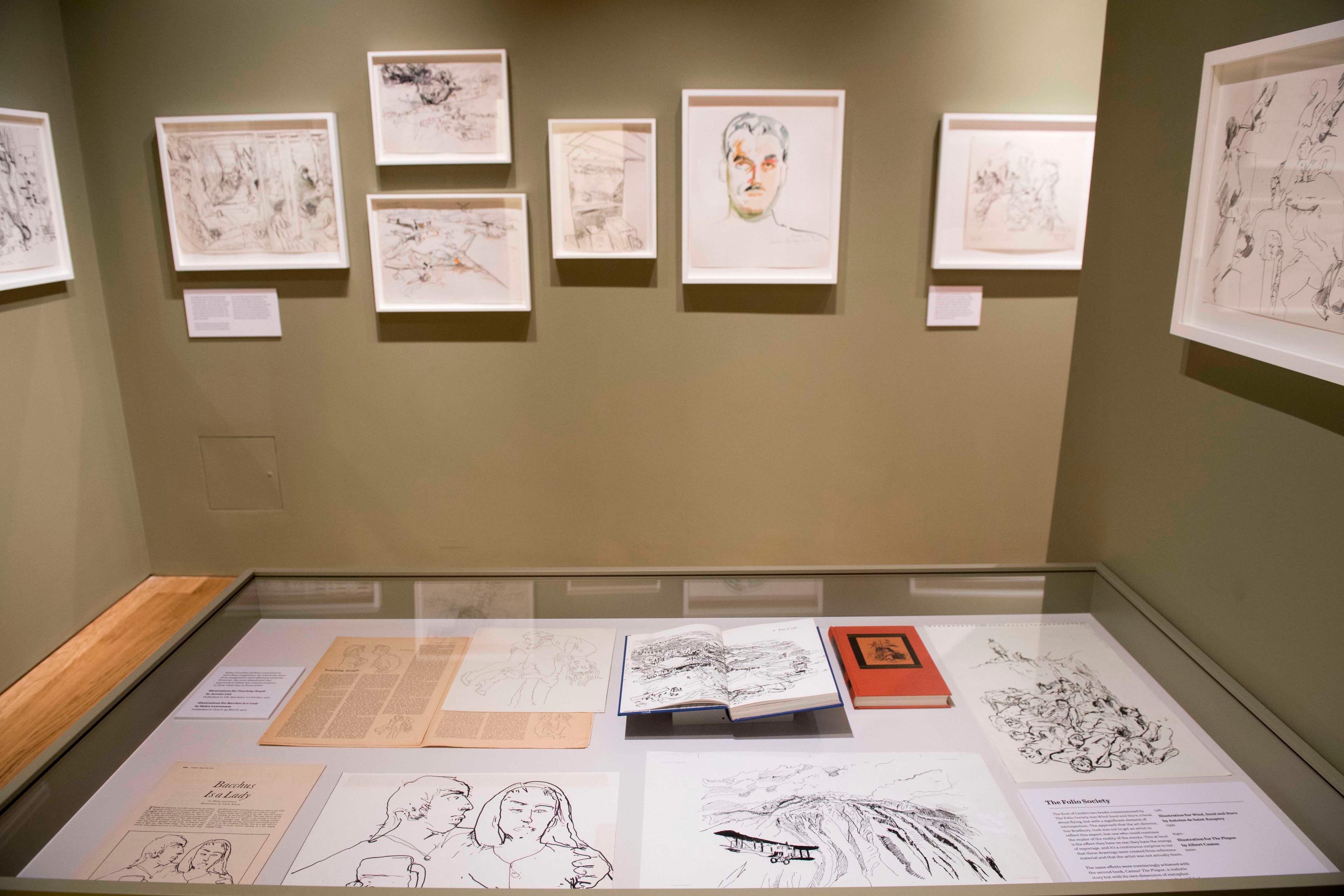 Framed illustrations displayed on a green wall with a glass display case showcasing additional illustrations in an exhibition space.