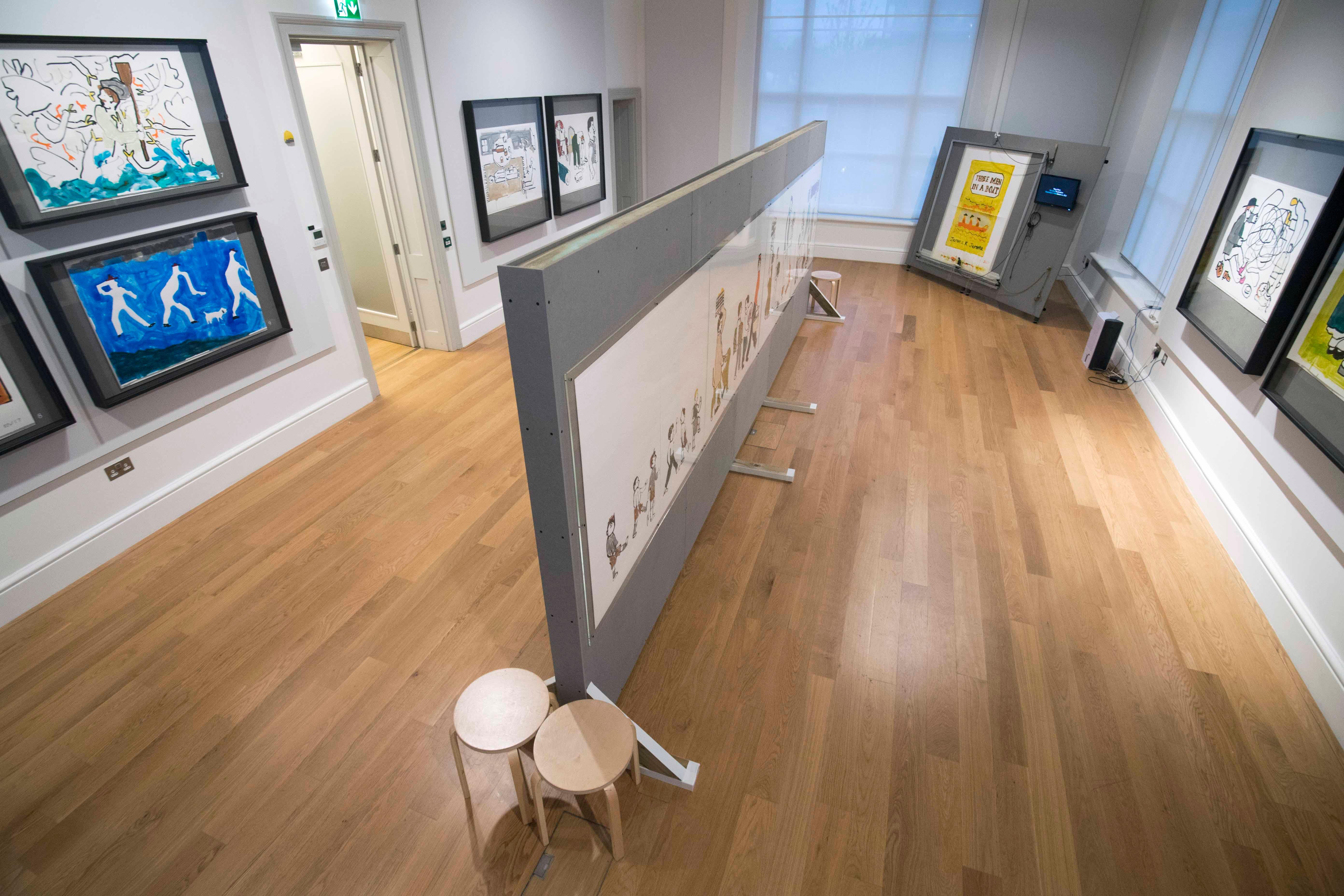 View looking down at an angle of an exhibition space with large illustrations on display, a large room divider is in the middle of the room with a large illustration of people on it.