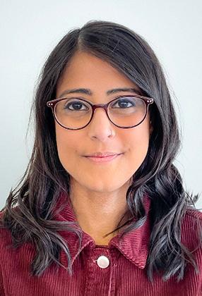A woman with glasses and dark hair wearing a maroon corduroy jacket, smiles softly against a plain light background.