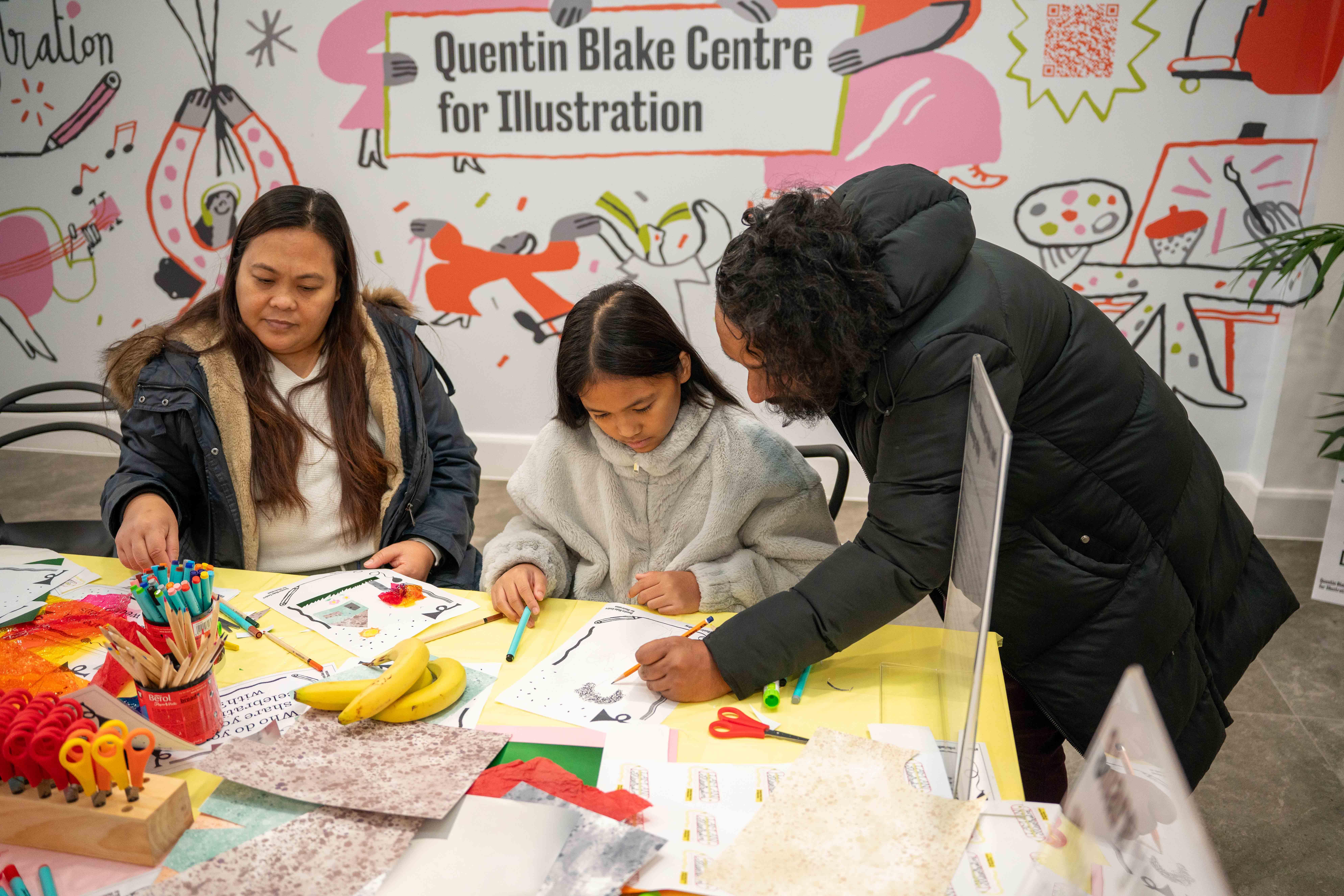 Photograph of three people, one is a child, at a table illustrating with pens and paper.