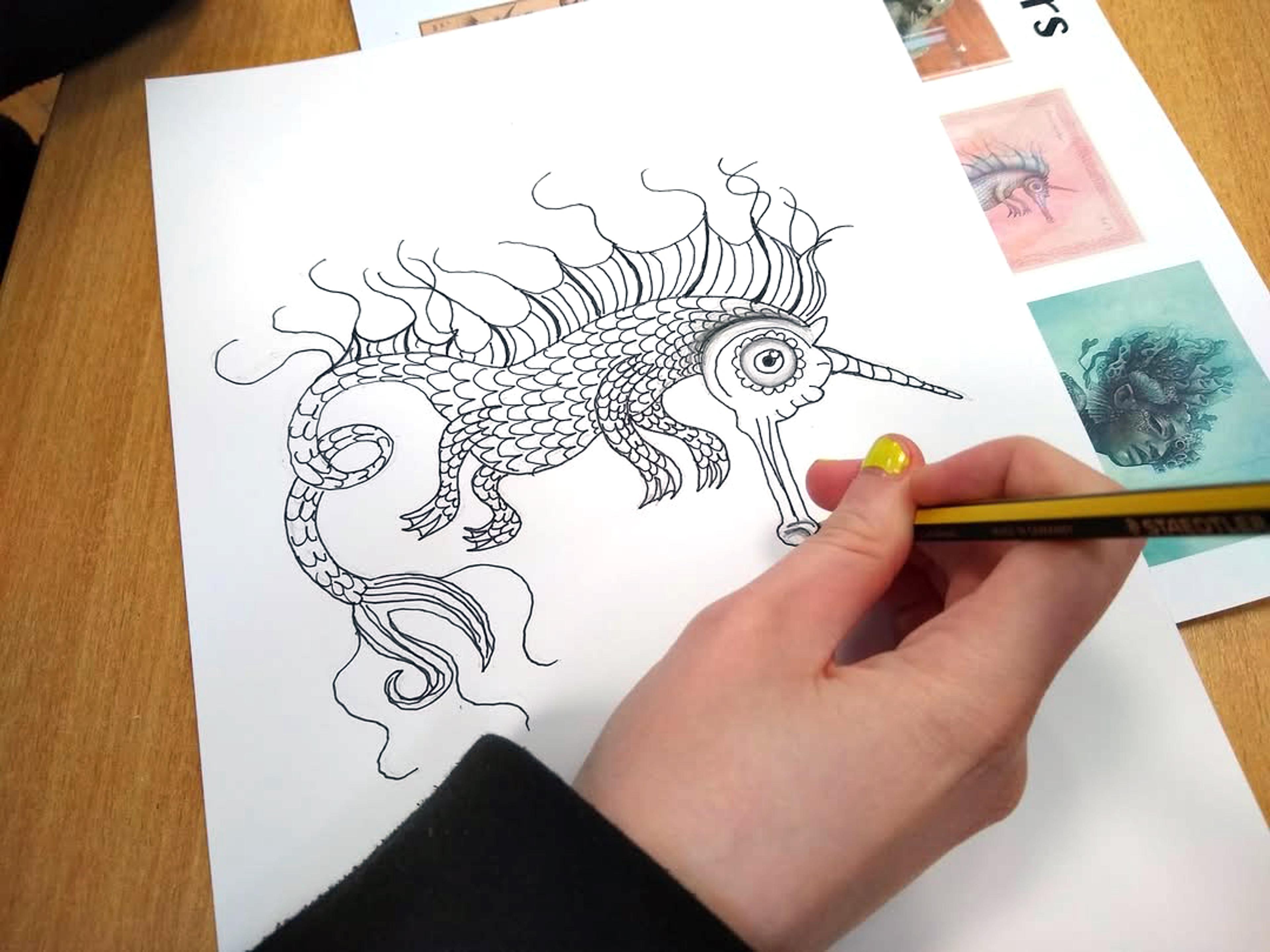 A photograph of a hand holding a pen, drawing a fantastical seahorse creature in black ink on a white piece of paper