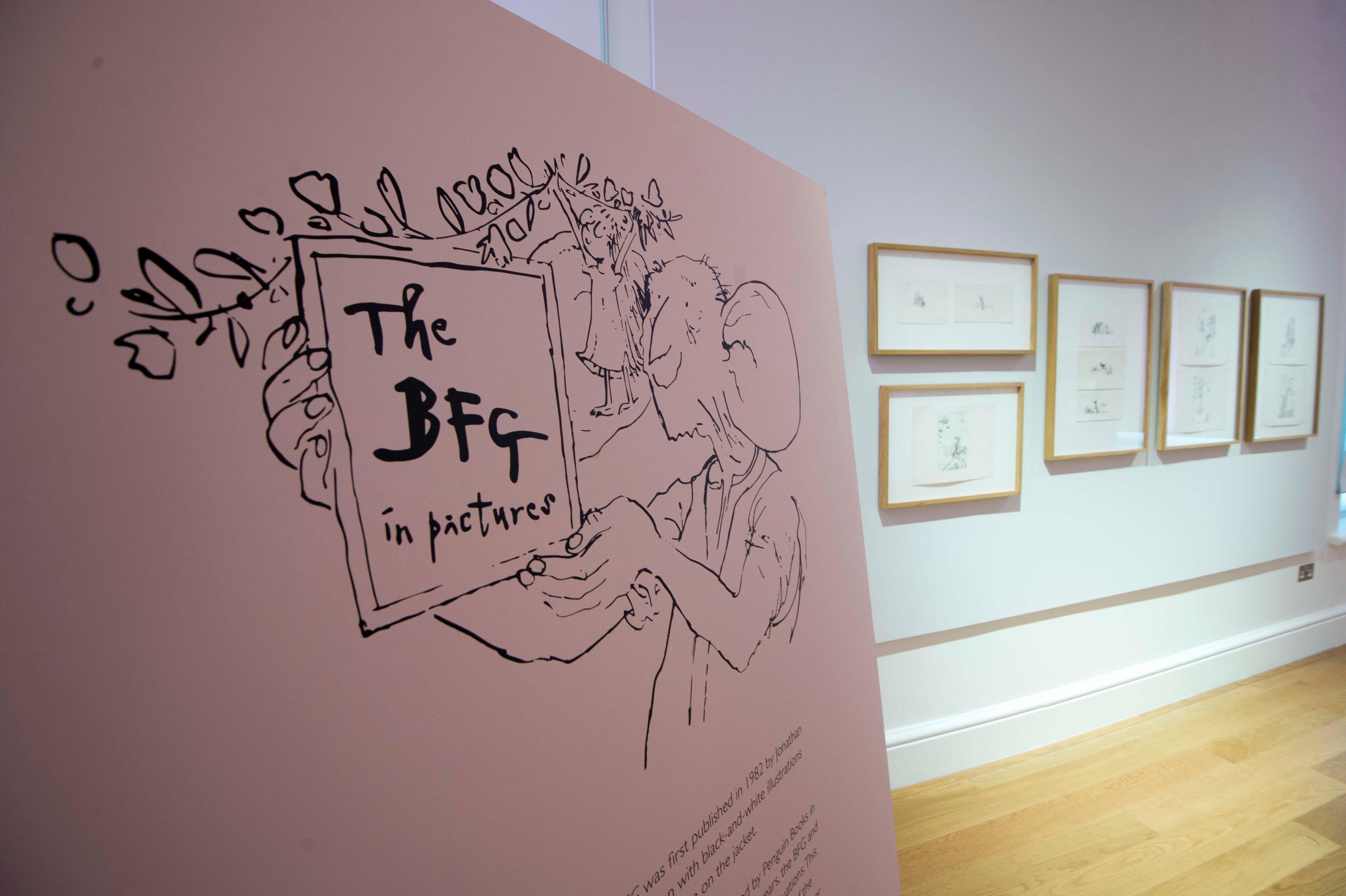 An illustration in an exhibition space with a character holding a sign with the text 'The BFG in Pictures', in the background are framed illustrations on the wall.