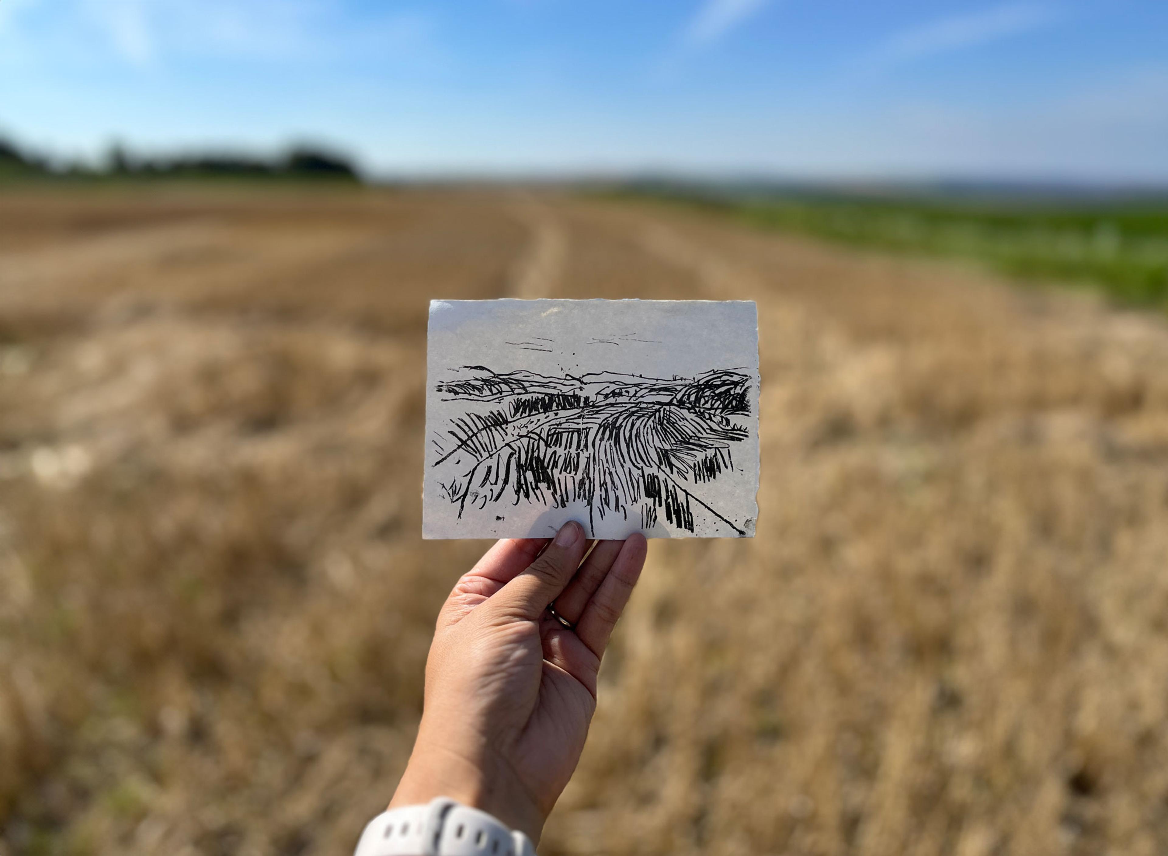 A hand holding a small landscape illustration on paper depicting  a field, behind it is a field and blue sky.