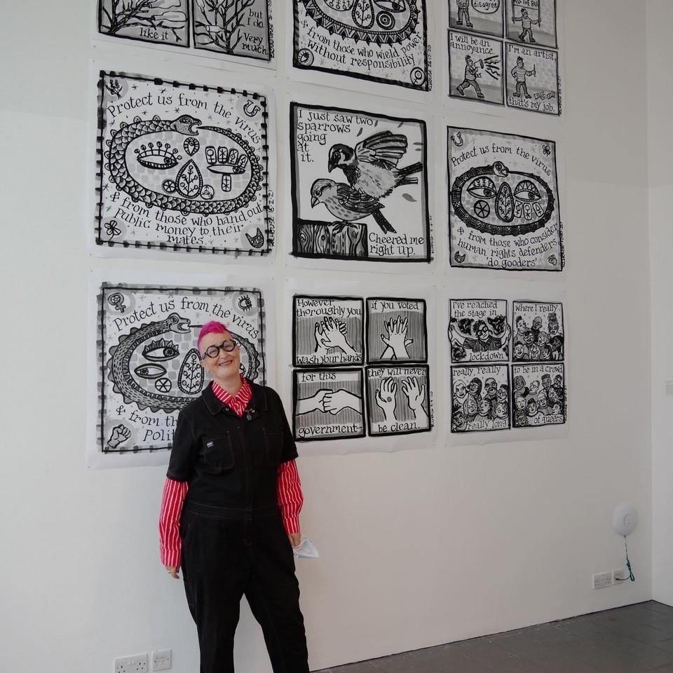 A woman with pink hair wearing a black jumpsuit over a red and pink striped shirt stands in front of an artwork display.