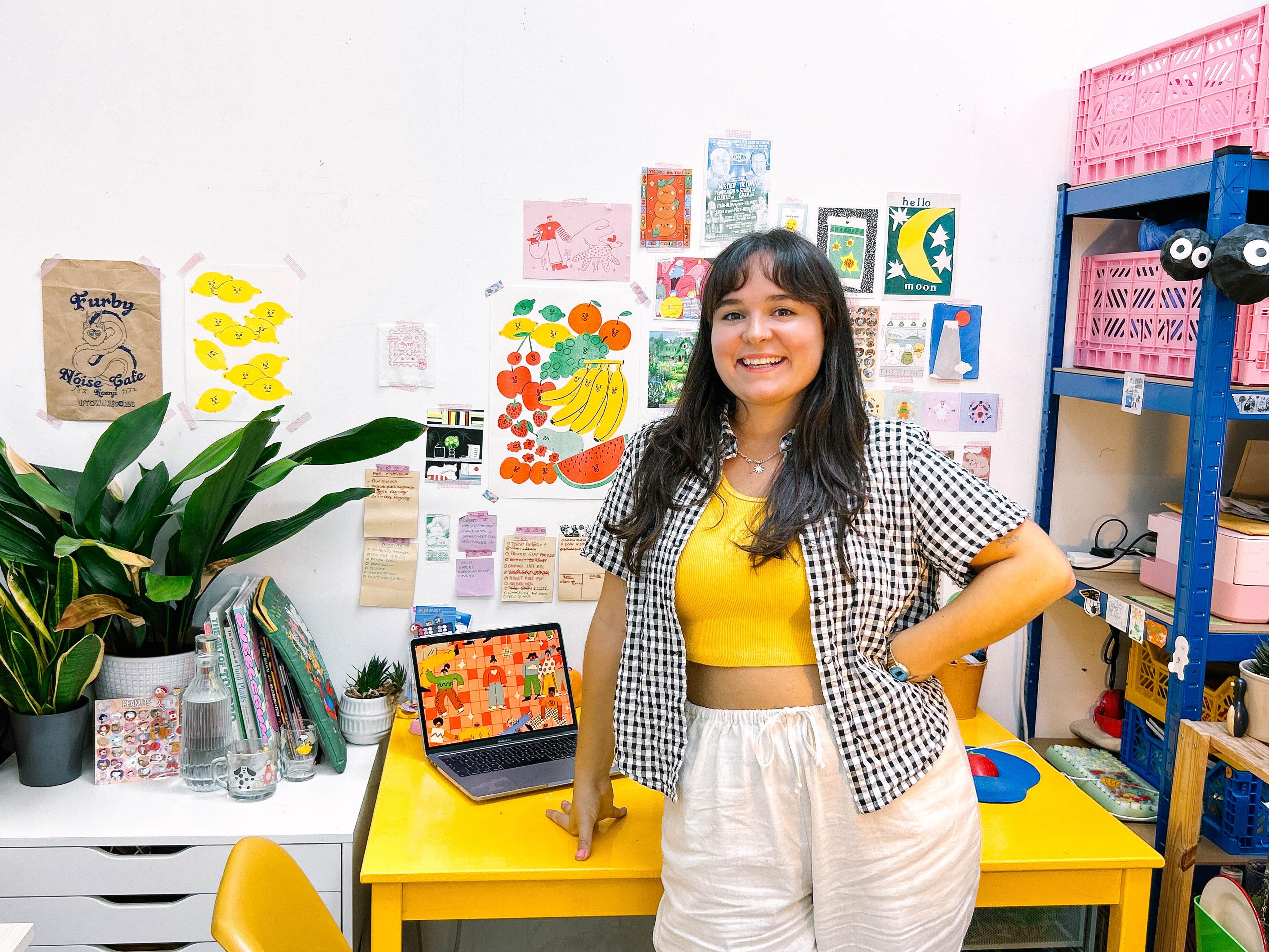 A person in a colourful studio space leaning on a desk looking ahead smiling. There are artworks stuck on the wall.