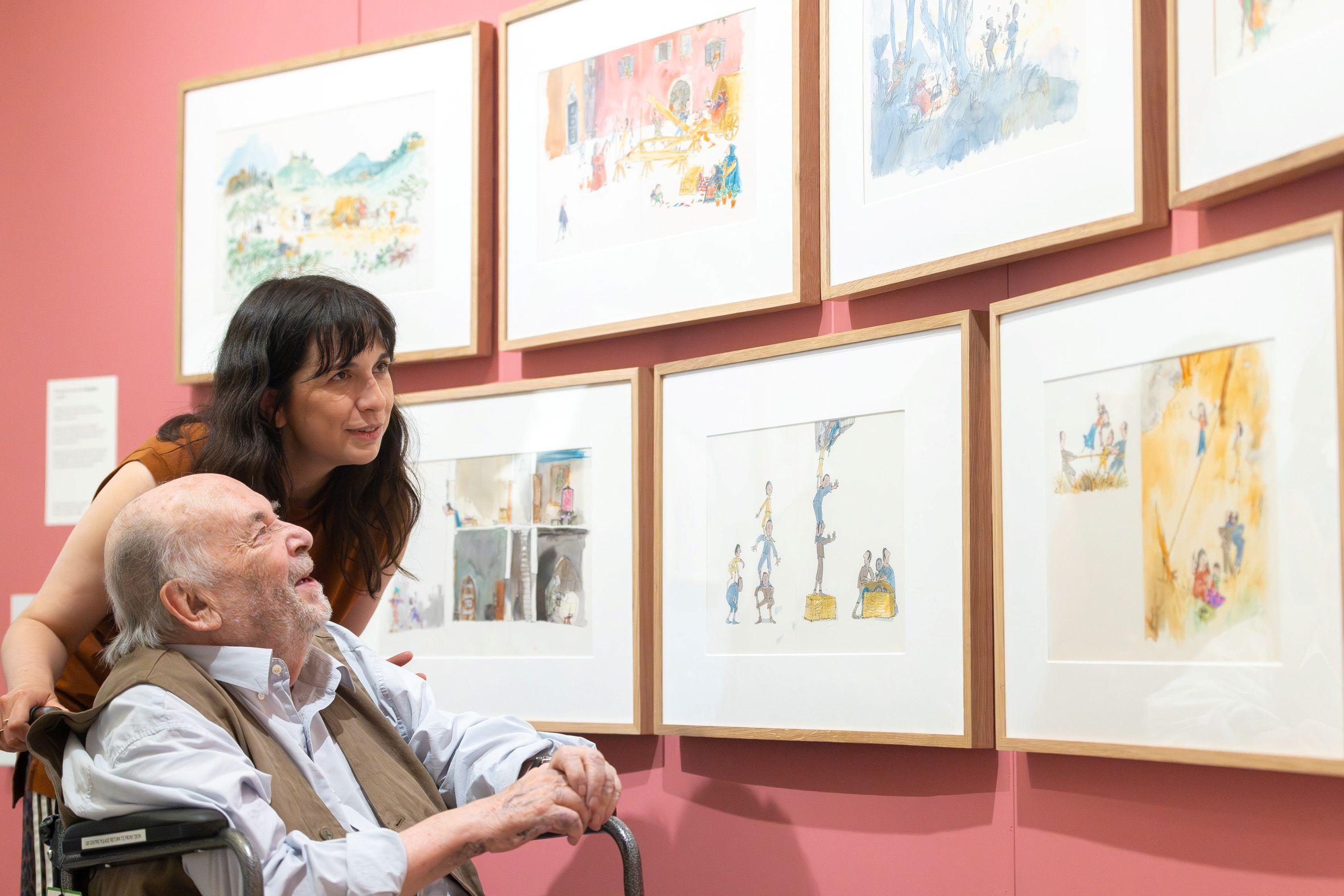 A friendly looking man in a wheelchair smiles up at colourful illustrations, accompanied by a woman with a fringe also looking up at the illustrations.