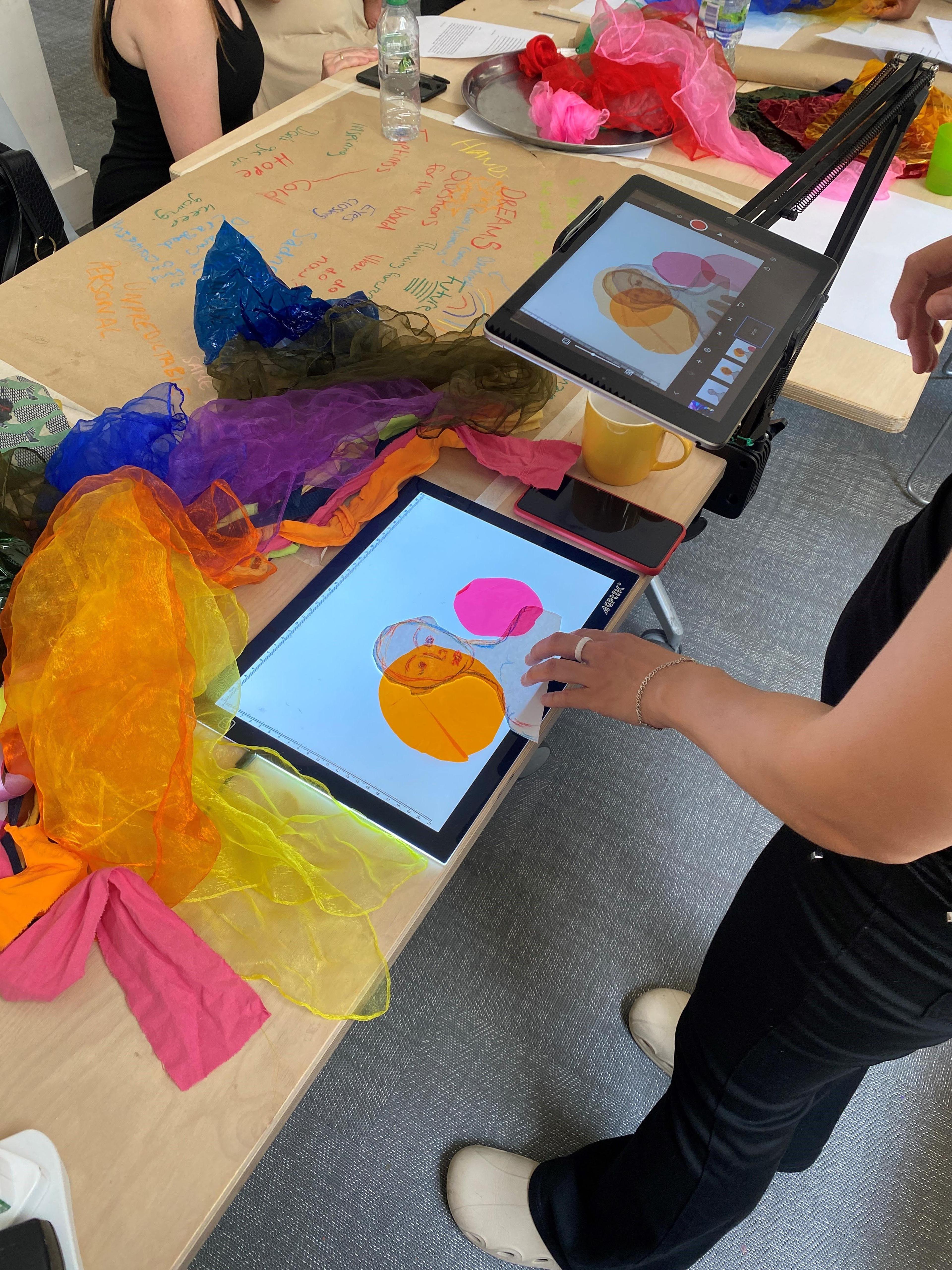 Photograph of a person creating an animation with a digital tablet on a stand, using sheets of fabric and a drawing of a person. On the table there is a pile of fabric and someone in the background also sitting at the table.