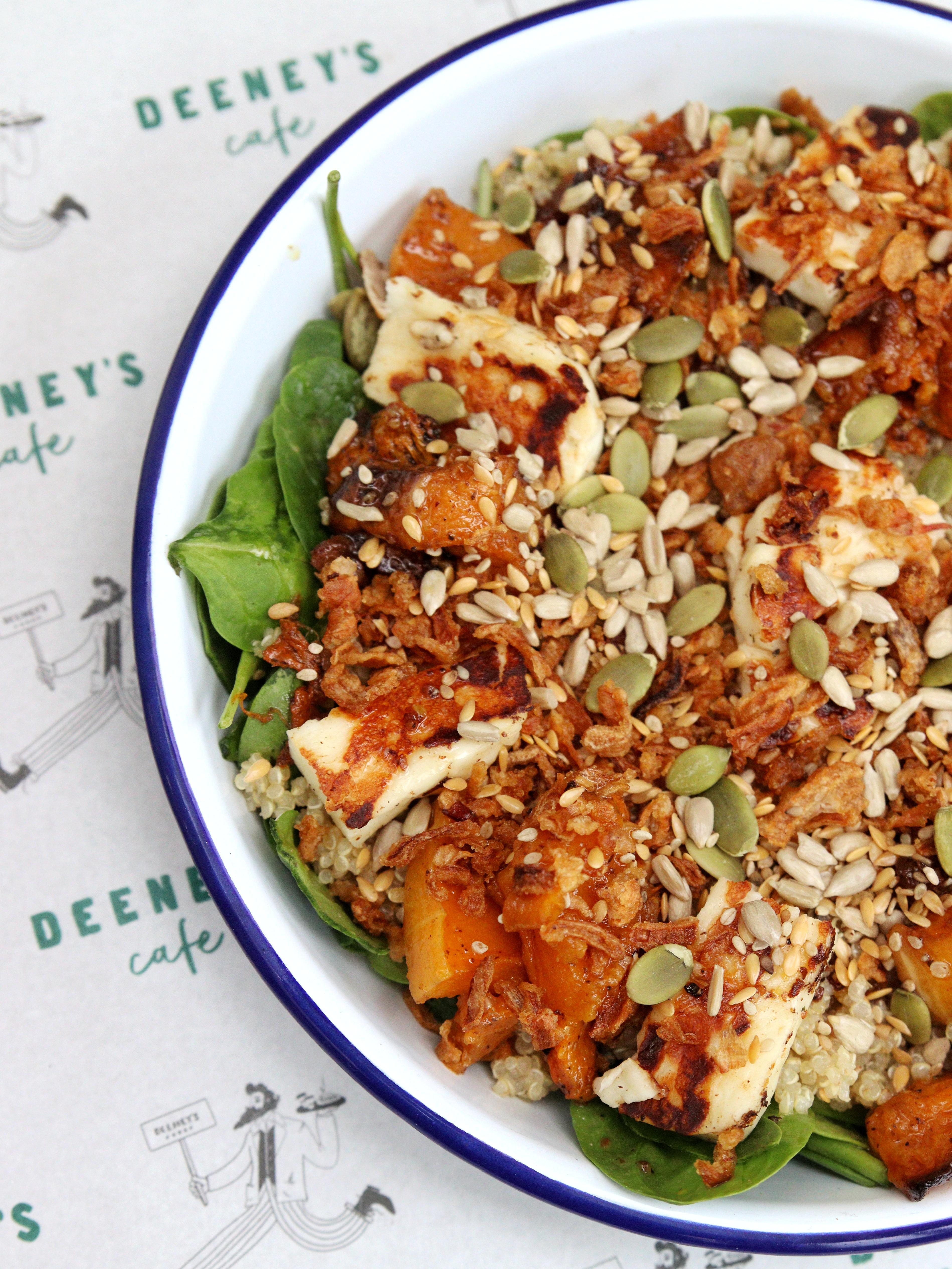  Close-up of a colourful salad topped with seeds on a blue-rimmed plate, against paper table lay labelled Deeney's.