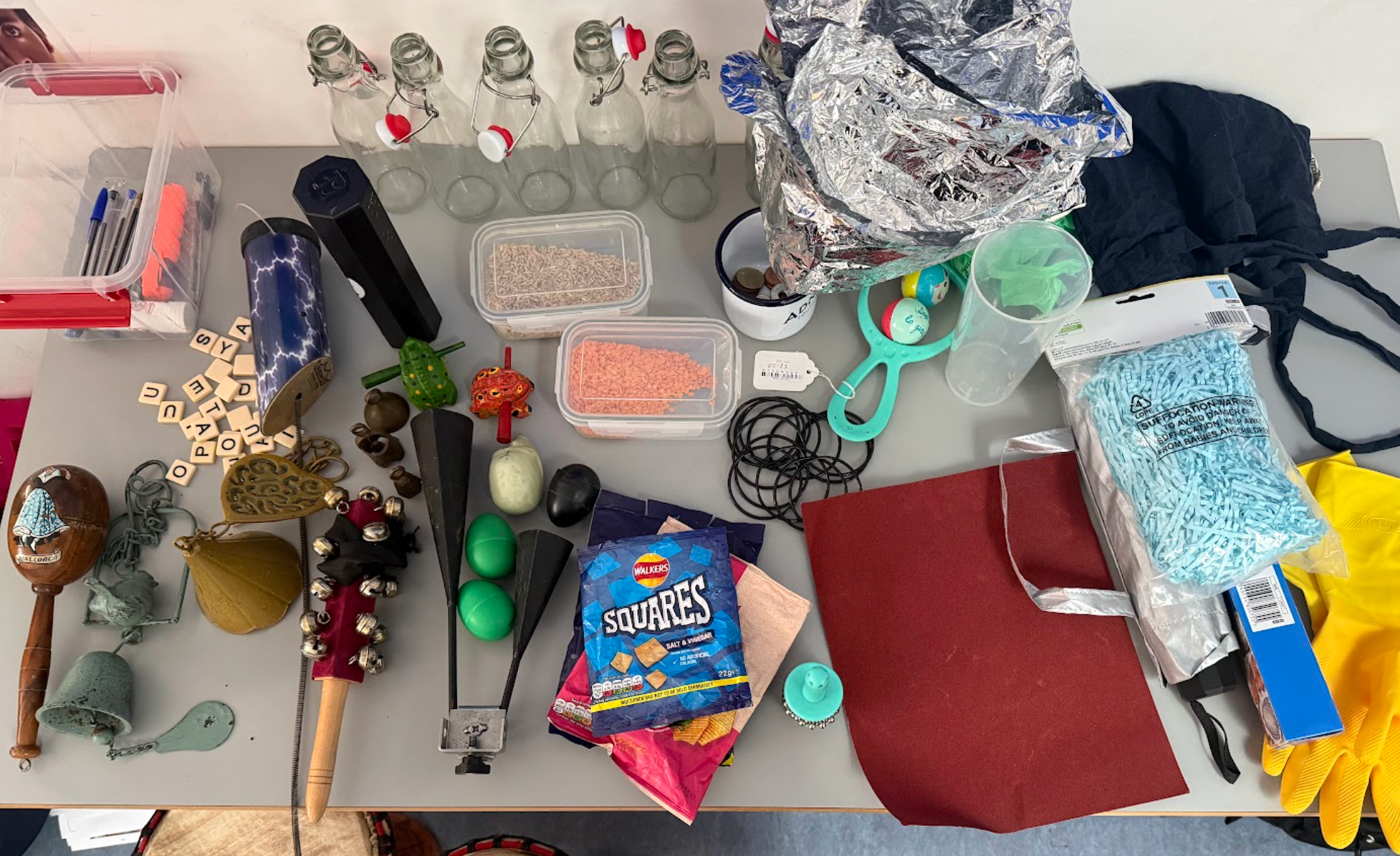 A table laid out with an array of everyday objects including glass bottles, maracas, scrabble tiles, crisp packets and lentils in tupperware.  