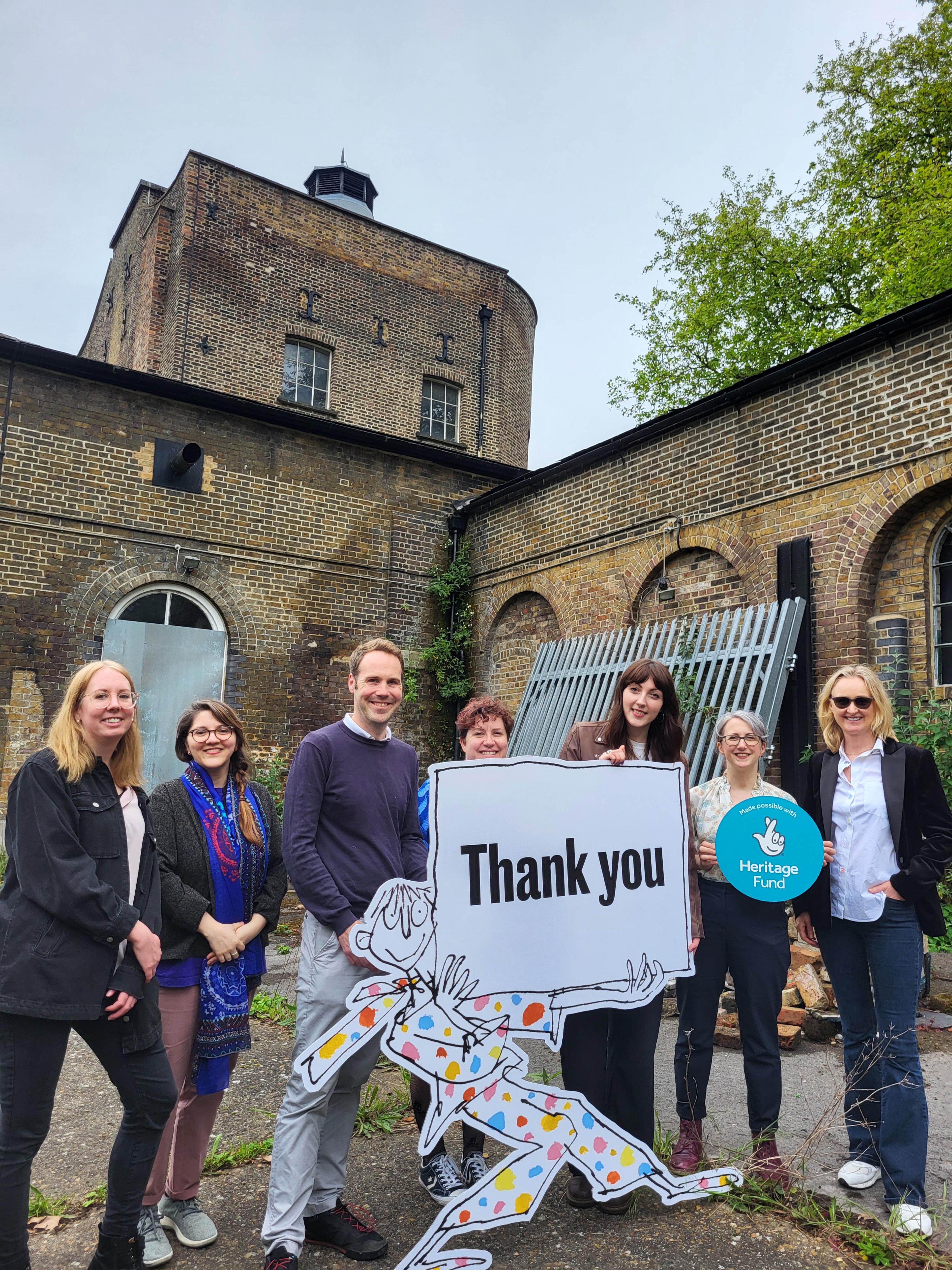 Photo of seven people standing in front of a large brick building holding a National Lottery Heritage Fund logo sign and a large illustration of a person holding a thank you sign.