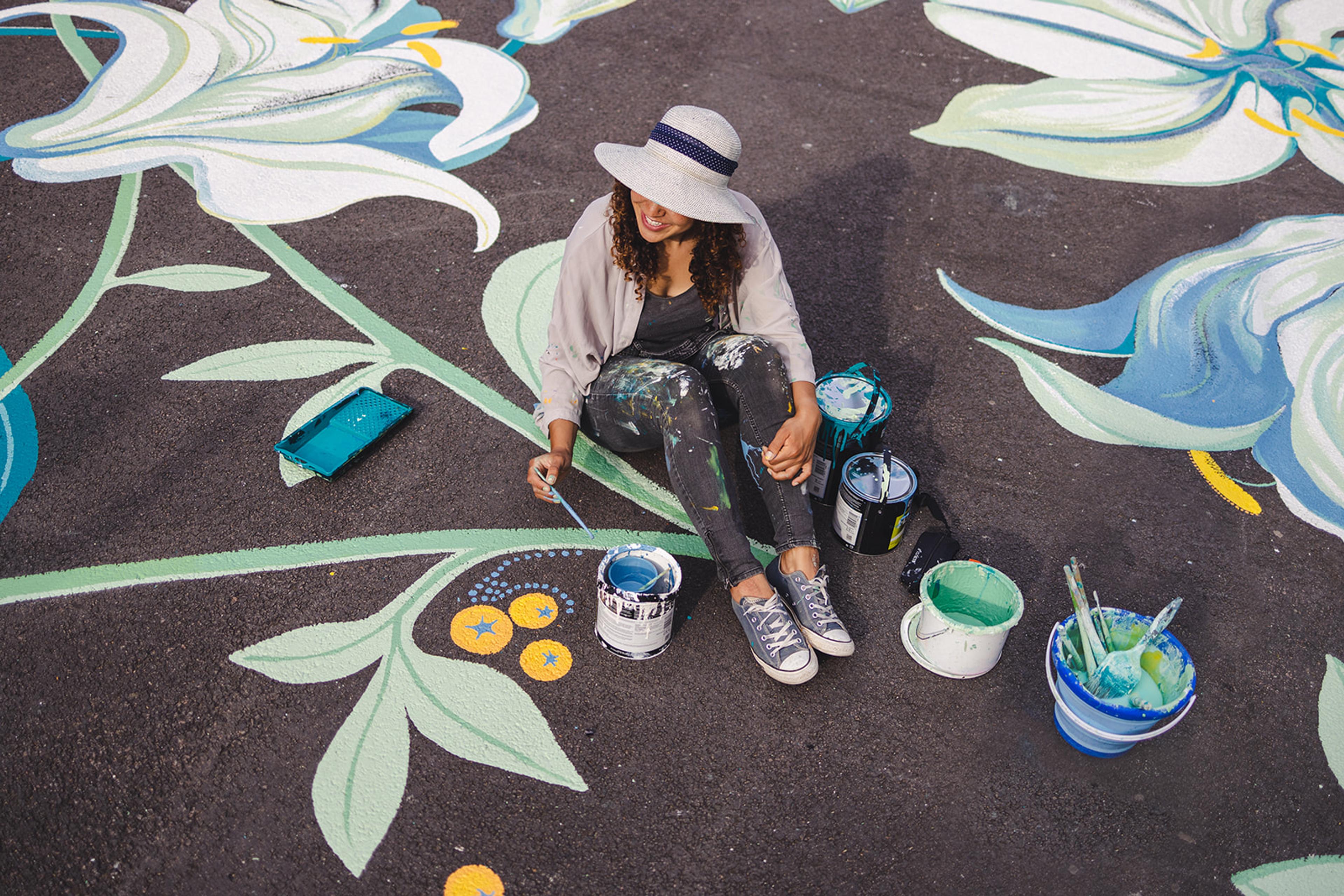 Photograph of person sitting on a street painting beautiful flowers and leaves all over the ground.