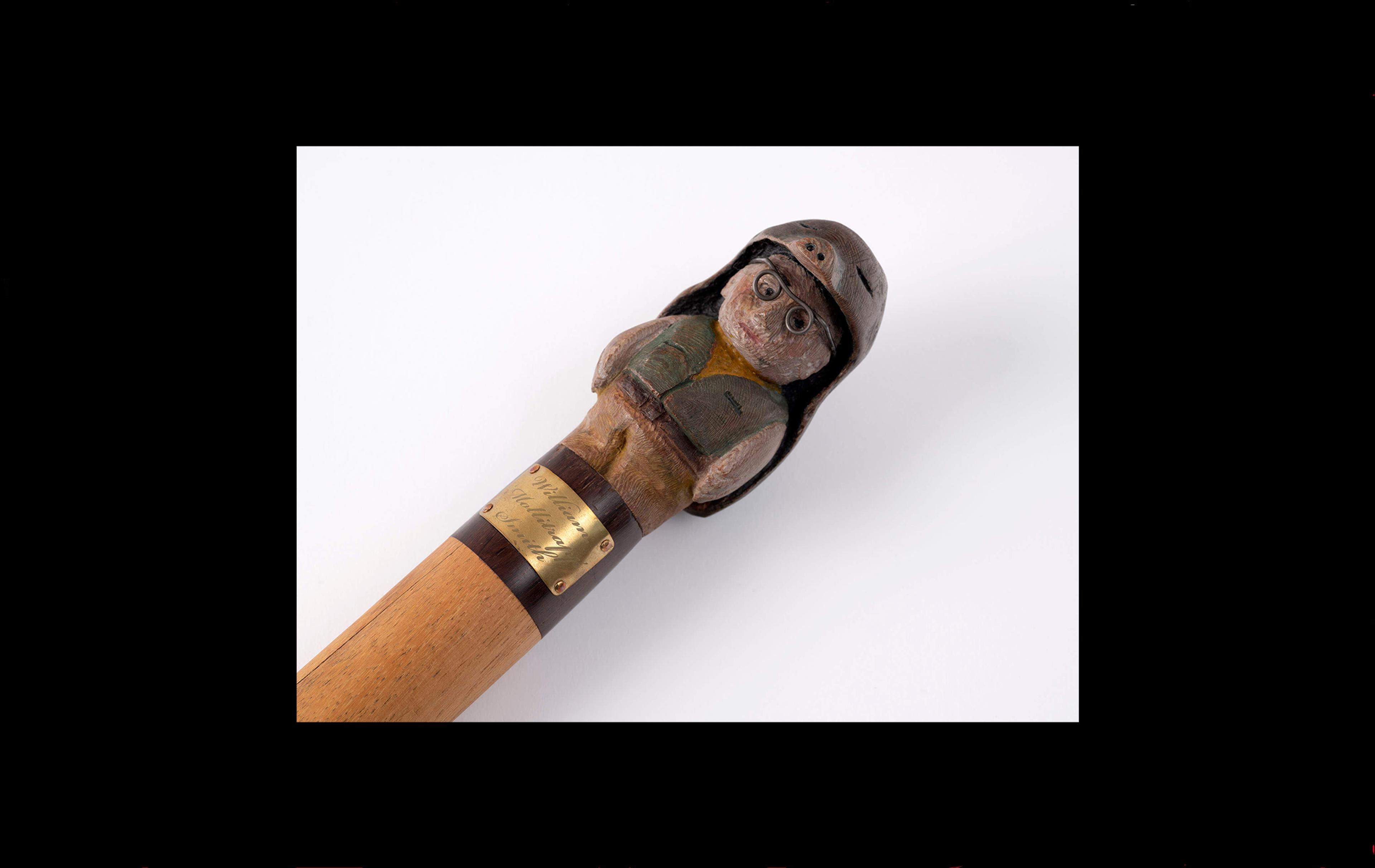 Photograph of a wooden staff with a carved figure at one end: a man wearing a giant moleskin as a costume