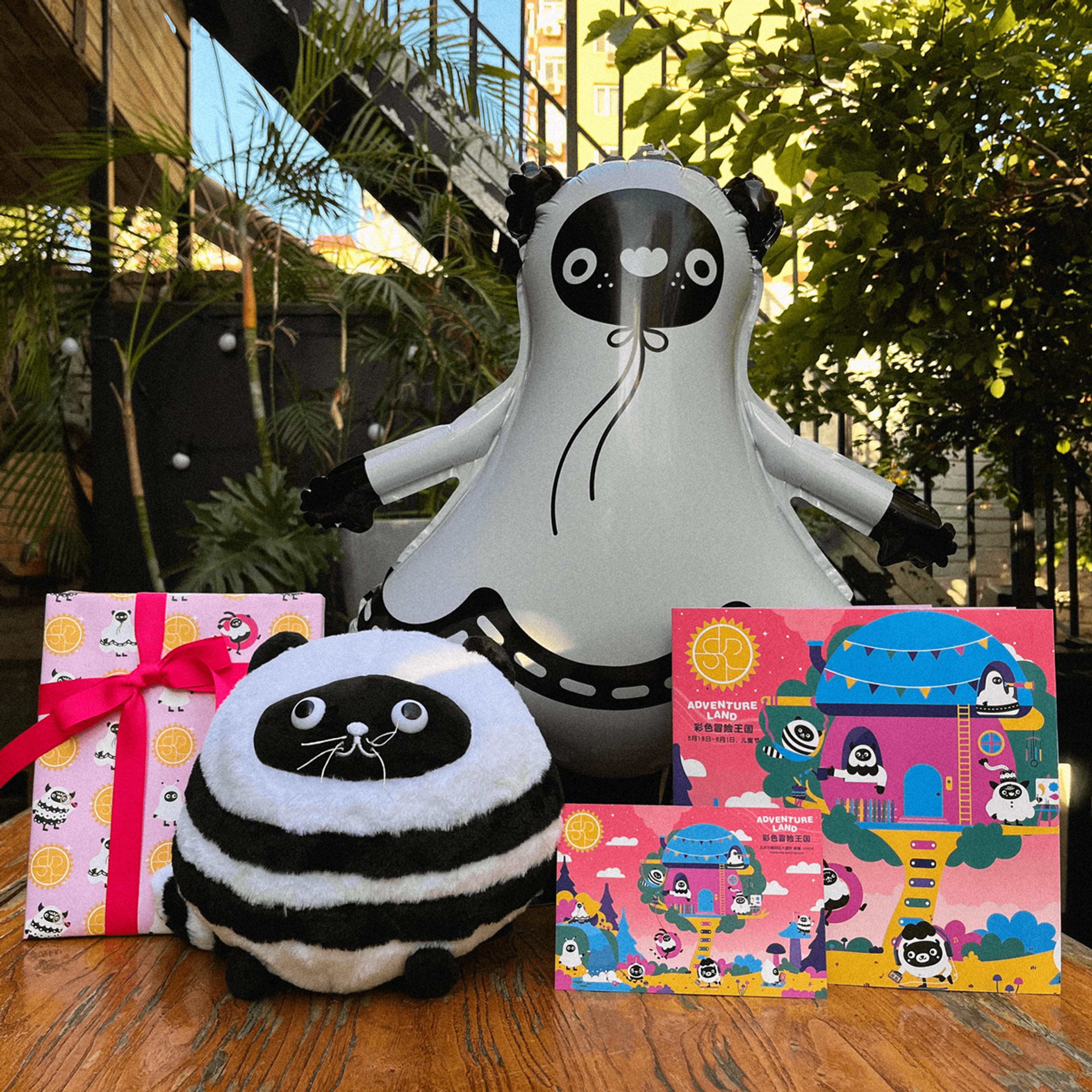 A photograph of a balloon and a stuffed toy in the form of a black and white furry animal character. The balloon and toy are displayed on a table next to some printed illustrated materials featuring the same characters and the words "Adventure Land".