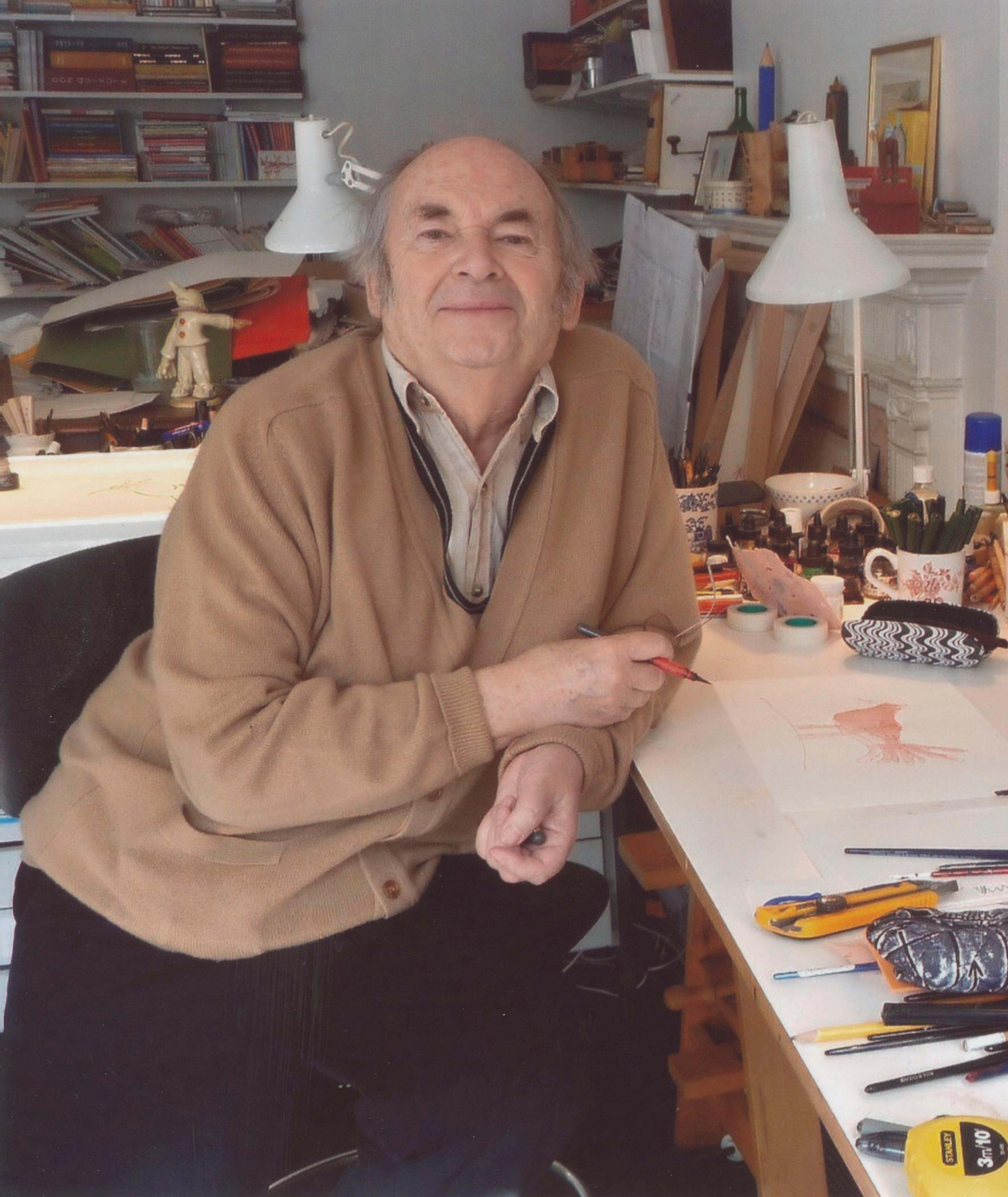 Quentin Blake sits at a desk, facing the camera, with a pen in his hand.