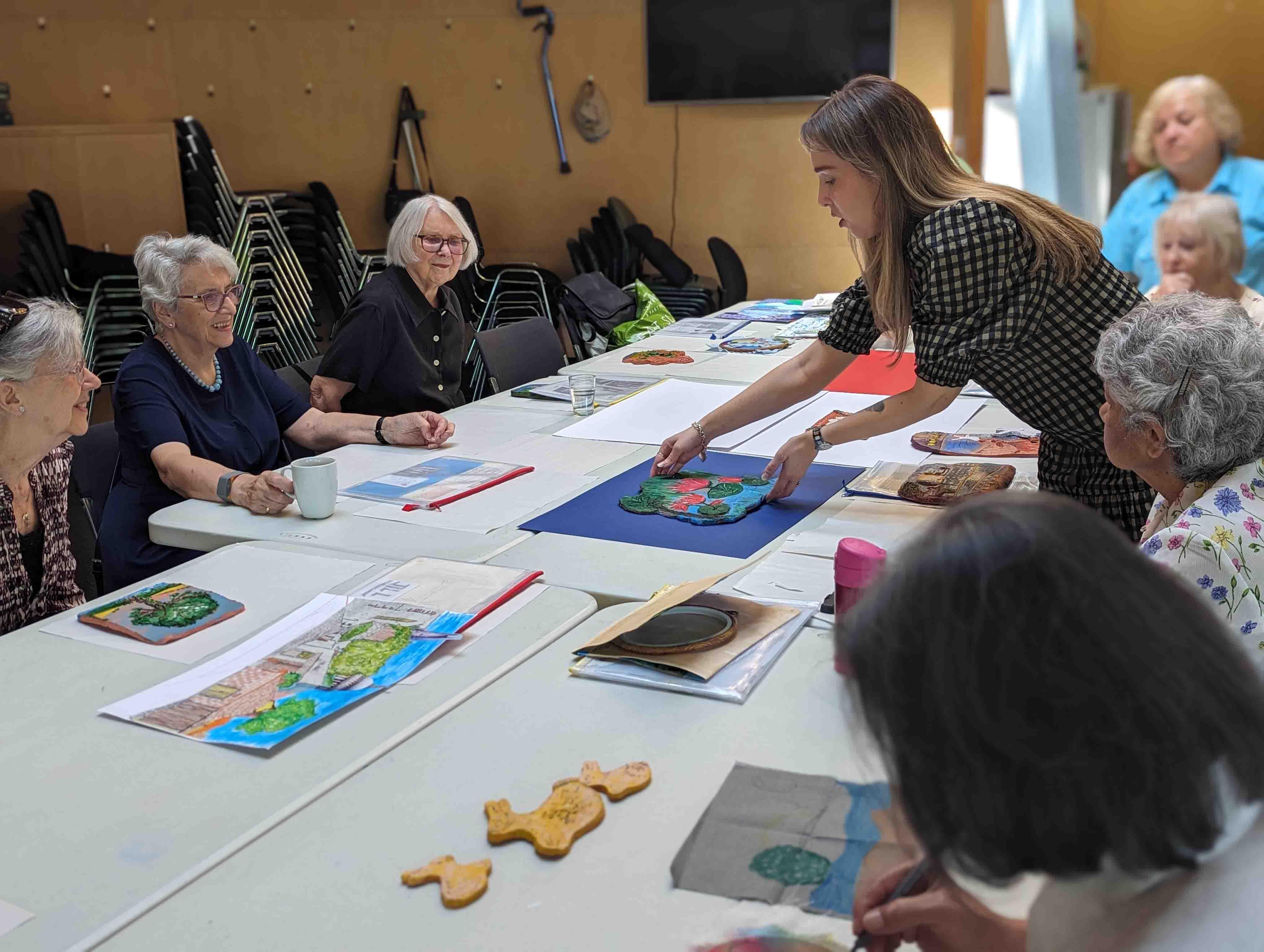Group of people sitting around a table painting ceramics. They are looking at a person standing up who is displayed a painted ceramic tile.