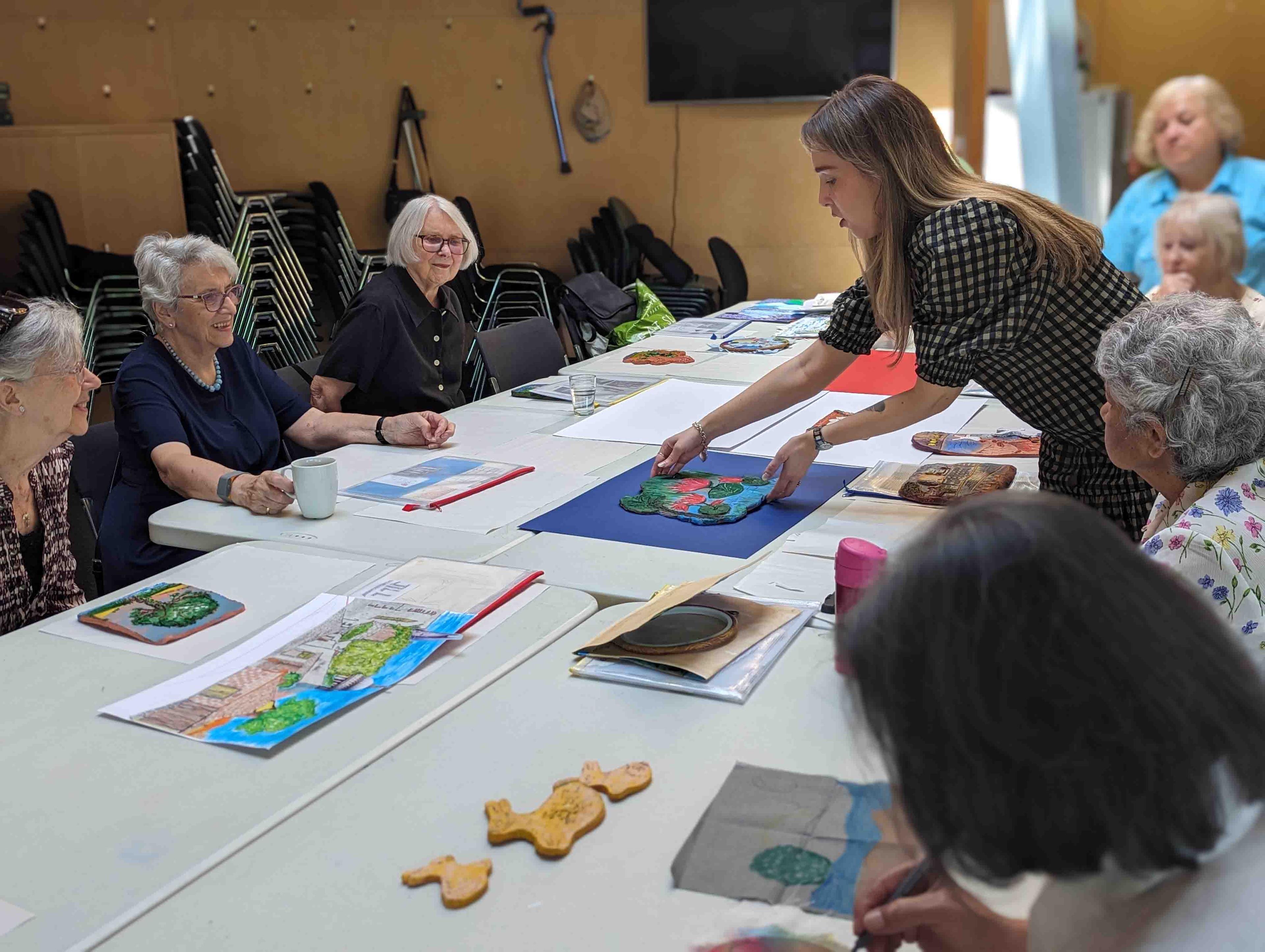 Group of people sitting around a table painting ceramics. They are looking at a person standing up who is displayed a painted ceramic tile.