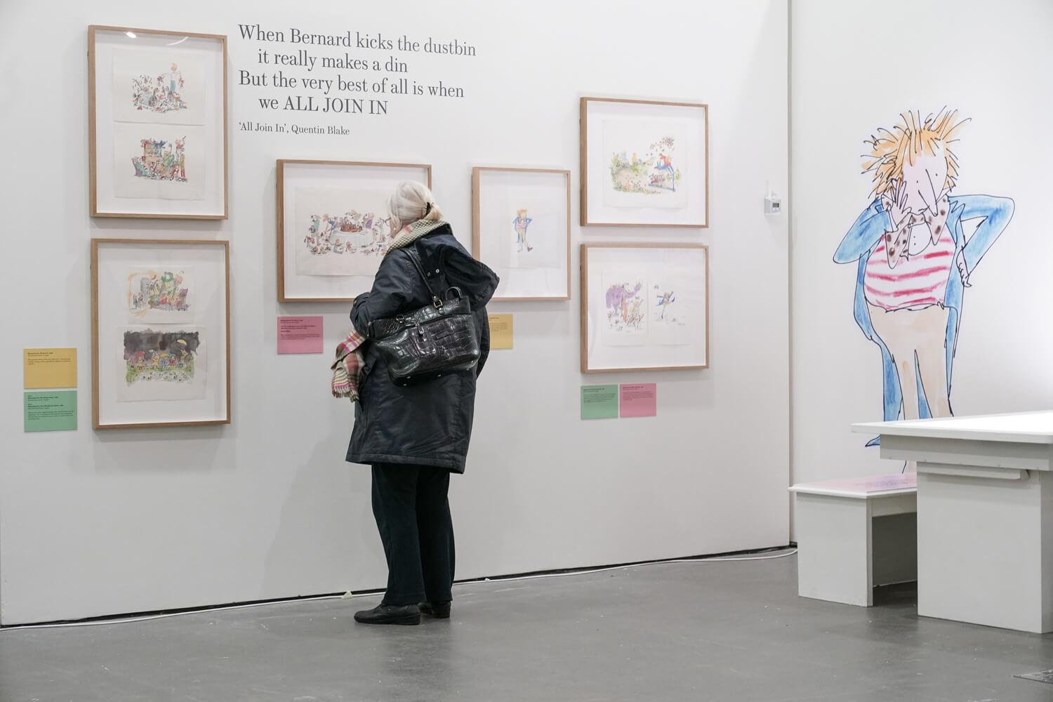 Person in an exhibition space with framed illustrations on the wall and a wall sticker of a person.