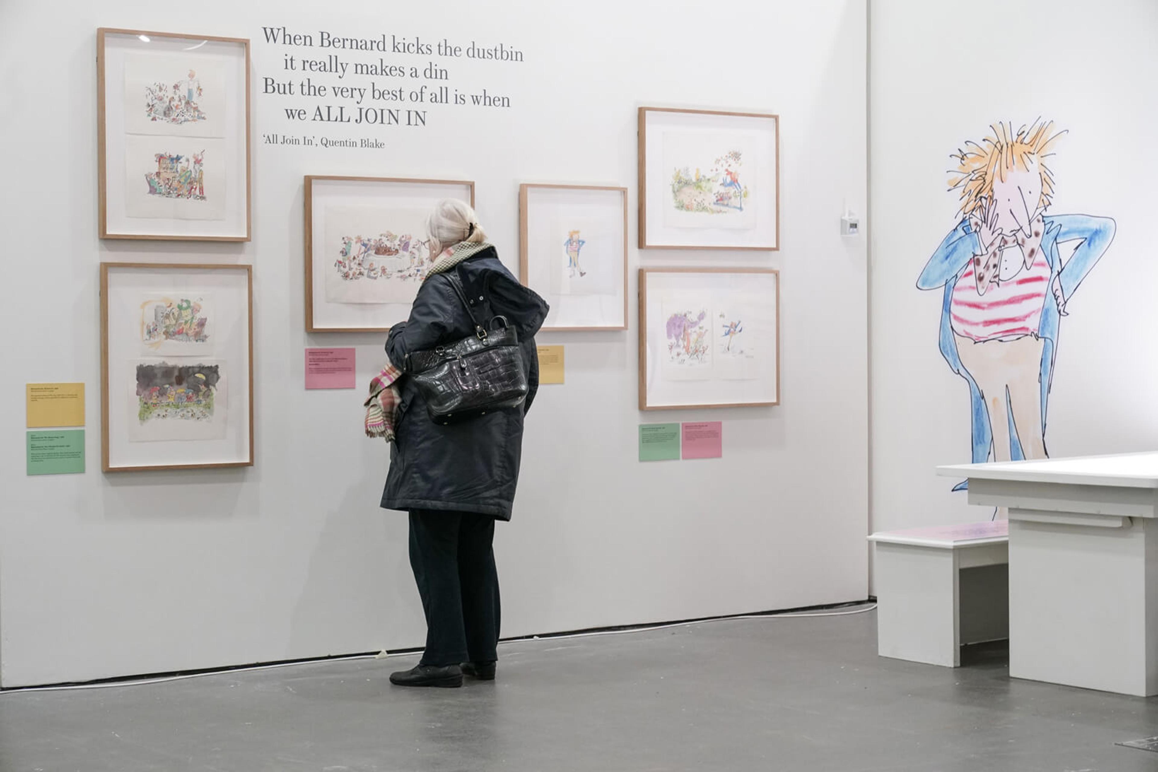 Person in an exhibition space with framed illustrations on the wall and a wall sticker of a person. 