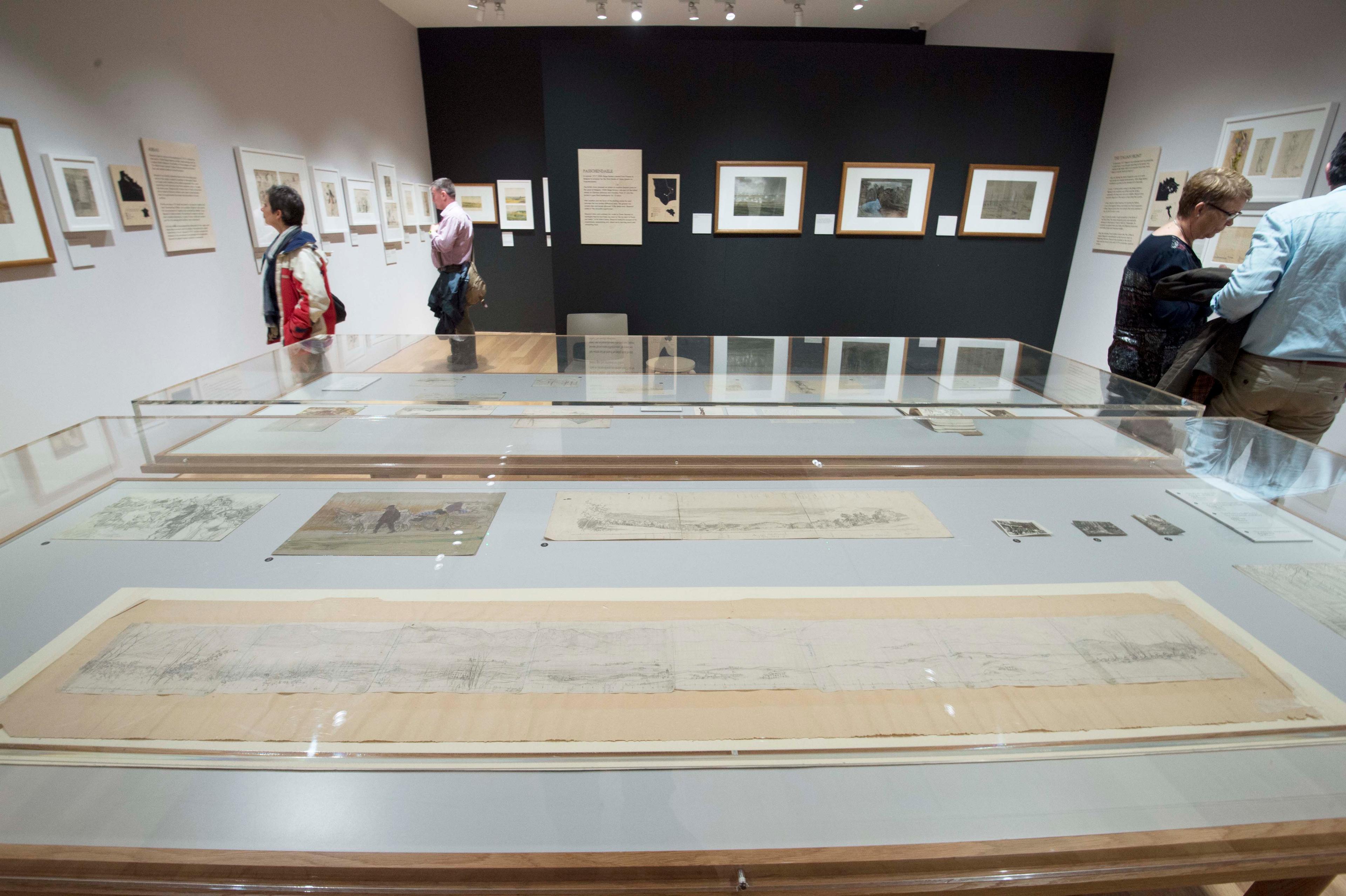 Inside of a gallery space with framed illustrations displayed on the wall. Two glass cabinets displaying illustrations are also in the space.