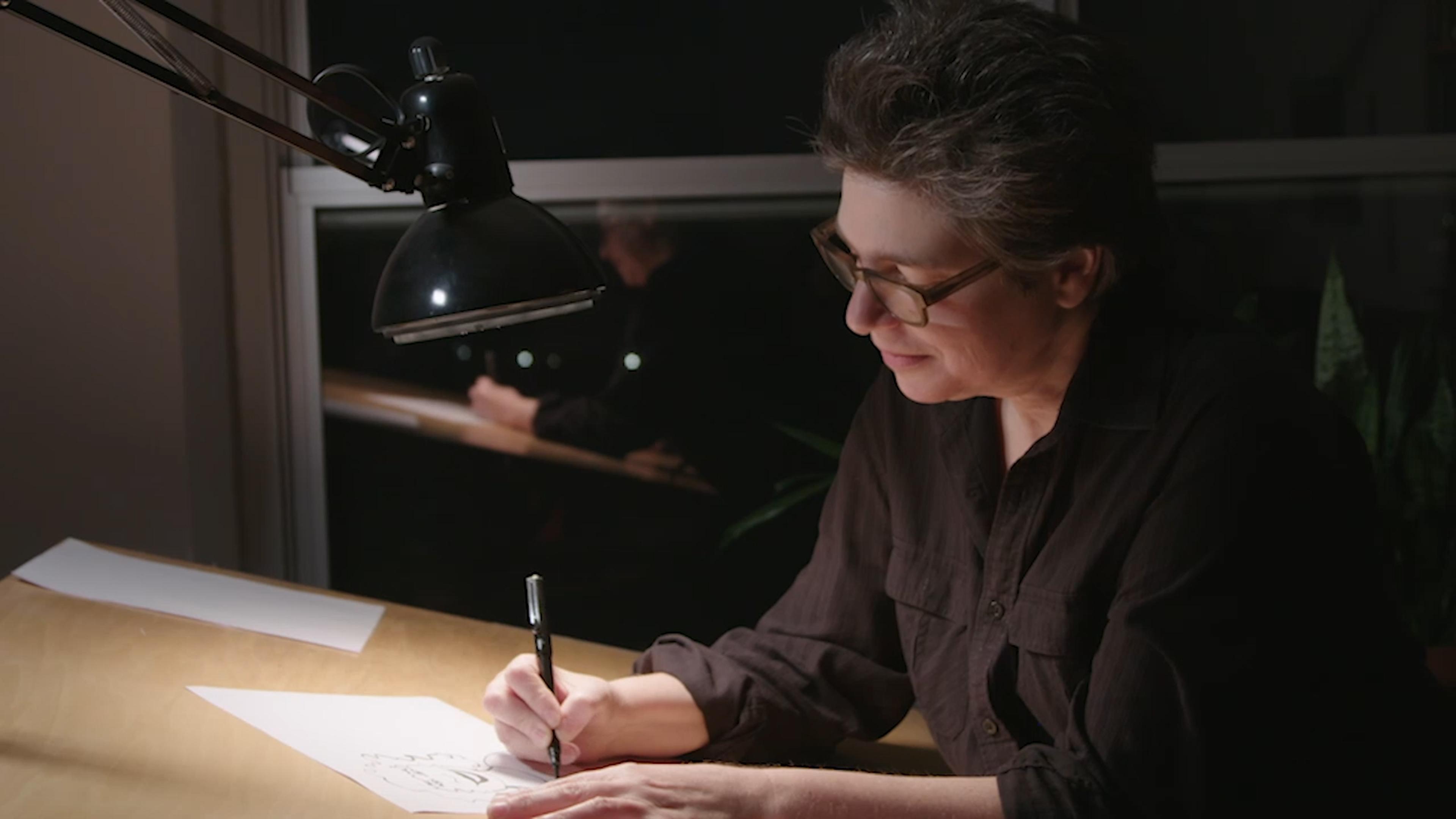 A woman sits drawing in a dark room with a small desk lamp shining on her work.