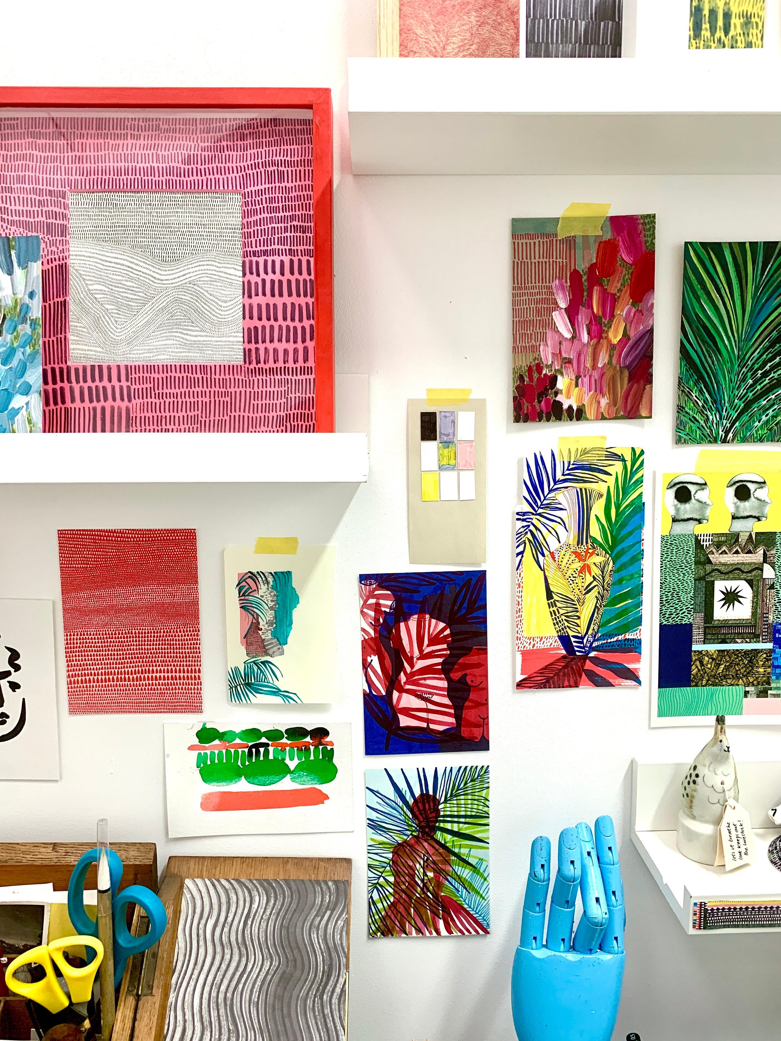 Photograph of lots of illustrations displayed on a wall and on some shelves. In front of the wall is a desk with a pot of illustrative tools and a model hand.