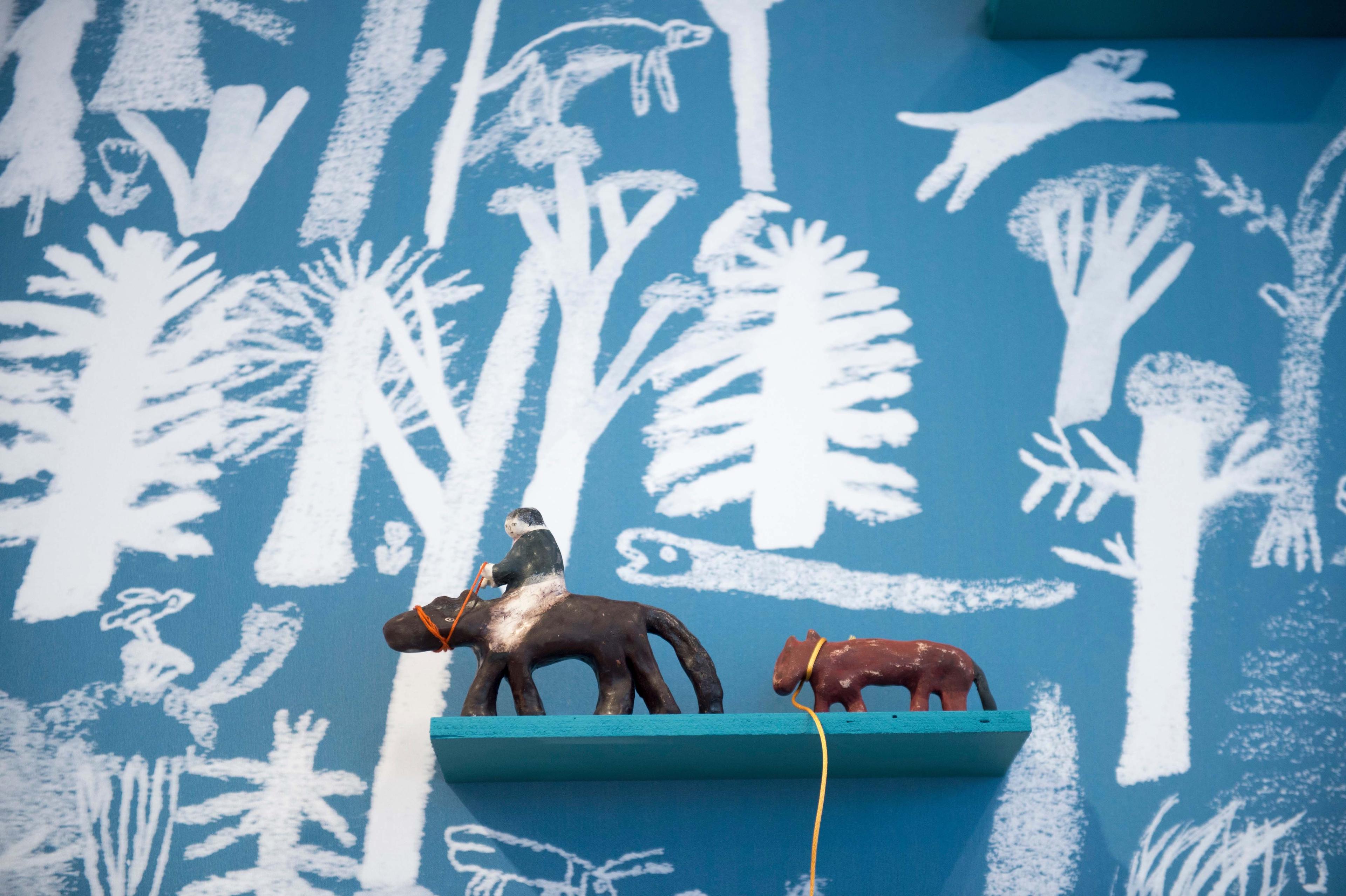 Two sculptures of two animals, a person is on the back of one of them. The wall behind is blue with an illustrated wood in white.